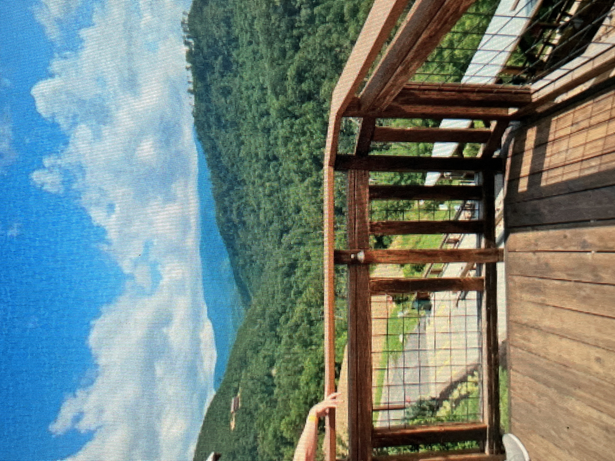 A high-angle, close-up shot captures a wooden deck with a scenic mountain view. The deck is constructed with wooden planks and features a wooden railing with vertical posts and a wire mesh infill. A person's arm and hand, adorned with a yellow wristband, are visible resting on the railing. In the background, lush green mountains covered in trees stretch into the distance, under a bright blue sky with scattered white clouds. The scene suggests an outdoor recreational area, potentially a viewpoint or a lodging with a view in Sejahtera, Indonesia.