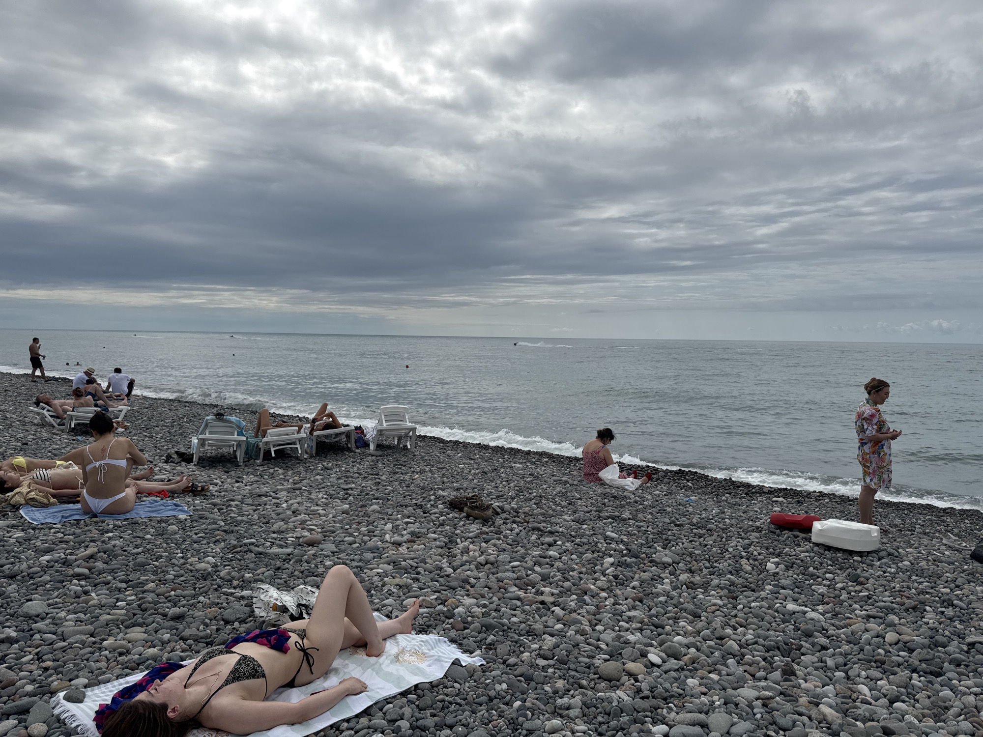 The image depicts a cloudy day at a pebbled beach in Batumi, Georgia. Several individuals are scattered across the beach, some lounging on beach chairs, while others recline on towels. In the foreground, a woman in a bikini lies on a towel, facing away from the camera. Further back, another woman in a floral dress stands near the water's edge. The sea is calm, with small waves lapping at the shore. The sky is overcast with heavy clouds. Scattered beach chairs and bags are visible amongst the pebbles.