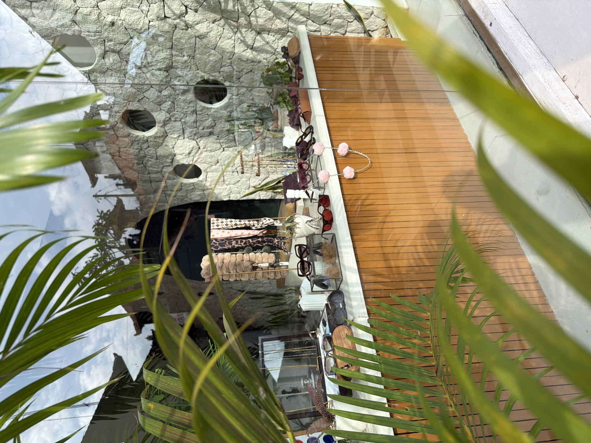 An upward-angled, candid shot captures a shop display window, viewed through dense, green palm fronds. The scene appears to be an outdoor or semi-outdoor setting in Kuta, Indonesia, given the tropical foliage and bright daylight.
The main subject is a merchandise display inside a glass window. A wooden slatted surface, likely a countertop or display table, holds an array of accessories. Prominently featured are numerous pairs of sunglasses, displayed on various stands and in clear cases. Some sunglasses have dark frames, while others show red or brown hues. Alongside these, necklaces and other jewelry items, possibly bracelets, are neatly arranged on stands. Other items include patterned scarves or bandanas, and furry, light-brown accessories, possibly hair ties or small bags. A distinct detail is a pair of pink pom-pom accessories, possibly keychains or bag charms, connected by a silver chain, hanging on the right side of the display.
Behind the wooden display table, the interior of the shop is visible, featuring a dark-colored mannequin dressed in a patterned garment. The back wall of the display area is made of a light-colored, textured stone with several circular openings, adding an architectural element. Reflections on the glass window obscure parts of the view, showing patches of a bright, cloudy sky and reflections of surrounding structures, indicating it is daytime with clear weather. Some indistinct text, possibly a brand name or product detail, can be vaguely seen on a dark item within the display, but it is not legible. The foreground is dominated by blurry, vibrant green palm leaves, partially framing the shop window and creating a sense of depth.