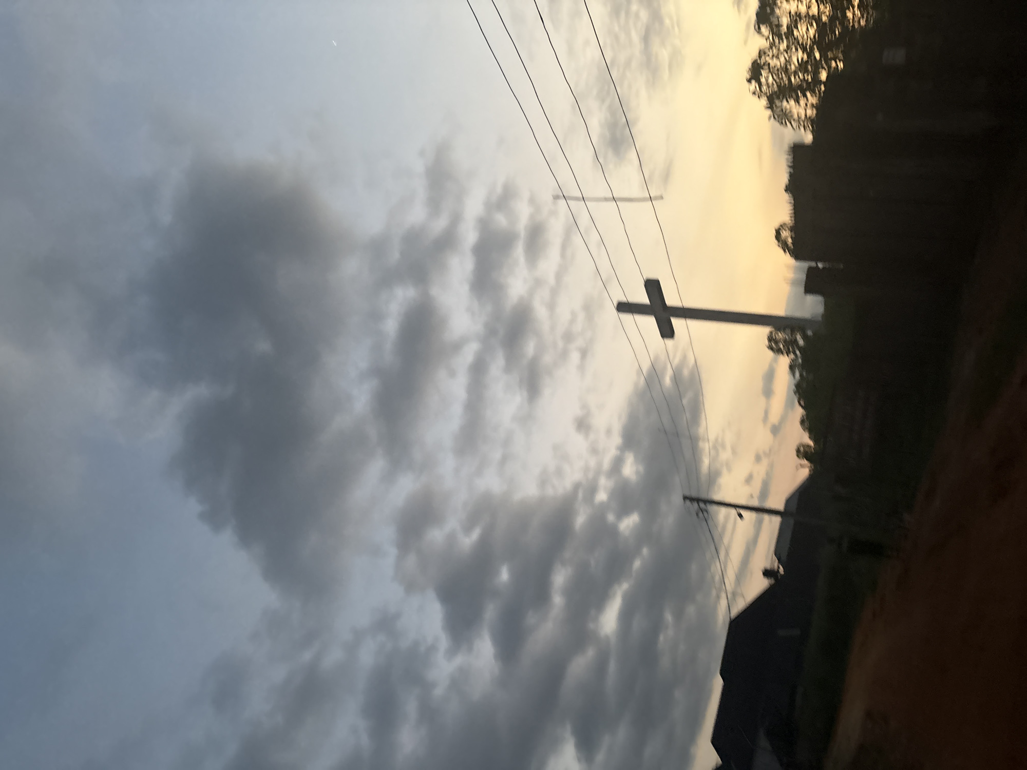 A low-angle, handheld shot captures a cross silhouetted against a cloudy sky at dusk in Obosi, Nigeria. The sky transitions from a pale blue at the top to a warm yellow-orange near the horizon. Dark clouds are scattered across the upper portion of the sky. In the foreground, a dirt road slopes down from left to right. To the left, the dark shapes of houses are visible. A utility pole with wires runs horizontally across the middle of the frame. A prominent cross stands to the right of center, casting a shadow. Further to the right, the silhouettes of trees and some unfinished structures are visible against the bright sky. The scene appears to be a residential area on the outskirts of the city.
