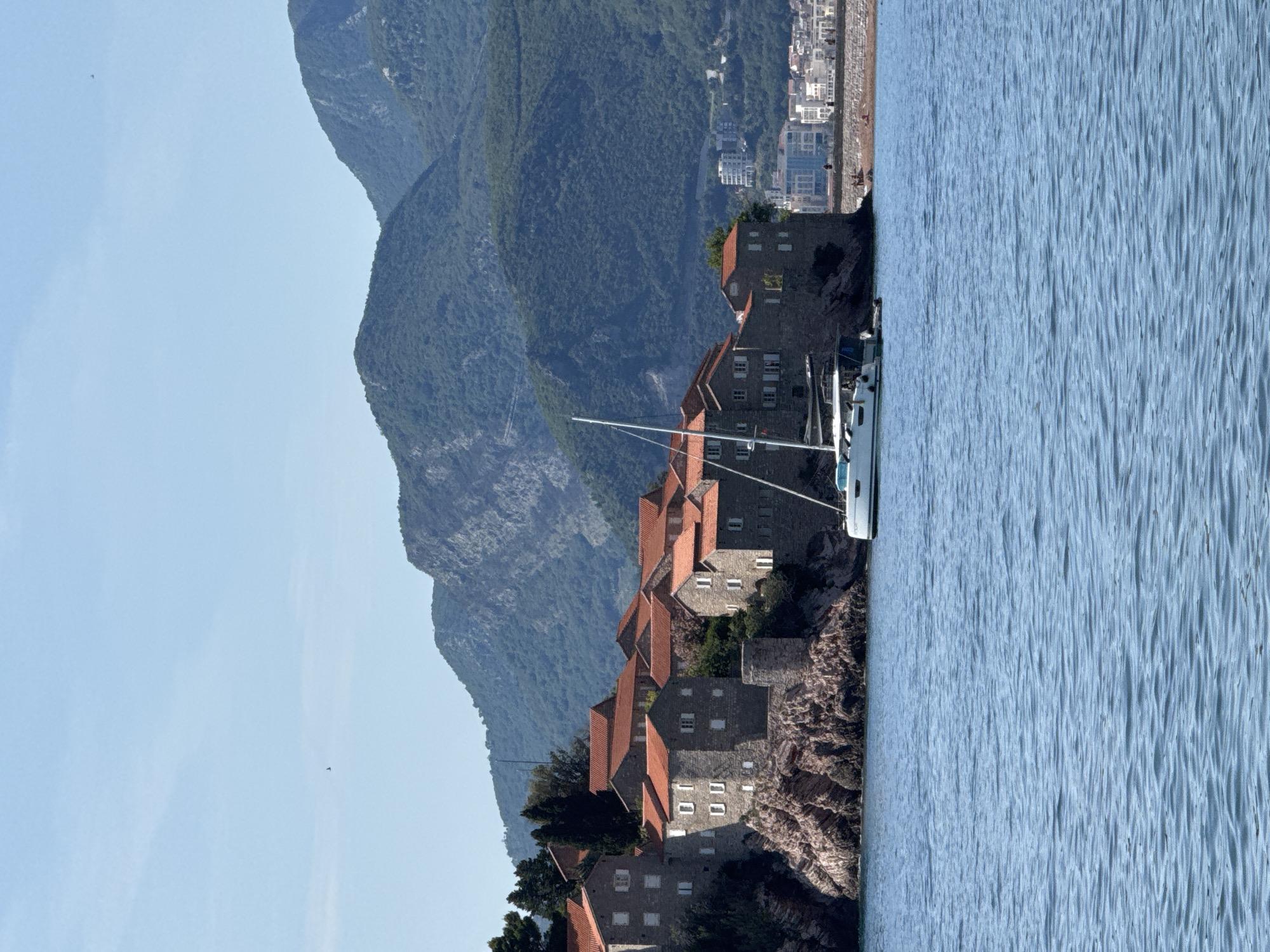 The image captures a panoramic view of Sveti Stefan, a fortified island village in Montenegro, under a clear blue sky with sparse clouds. The village, situated on a rocky islet, features numerous stone buildings with reddish-orange tiled roofs and white-shuttered windows, densely packed and appearing to be integrated with the natural rock formations. A white catamaran with its mast visible is anchored in the calm, blue-green water in the foreground, positioned to the right of the islet. In the background, majestic, tree-covered mountains rise steeply, dominating the horizon. To the far right, beyond the island, a modern coastal town with white and blue multistory buildings and a stretch of beach or promenade is visible, with tiny figures of people discernible on the shoreline. The overall scene depicts a serene coastal landscape with historical architecture contrasting with natural beauty and distant modern development.