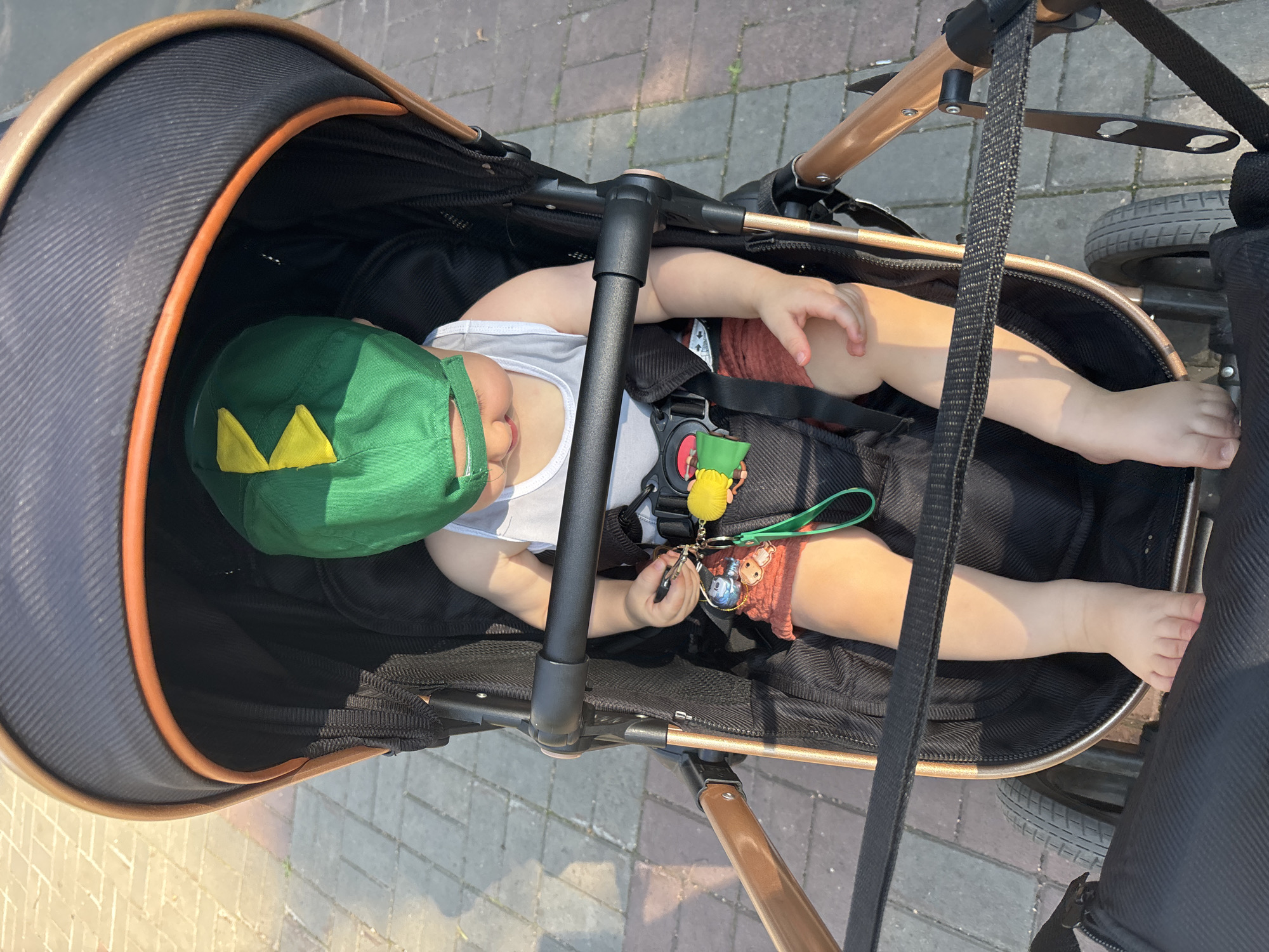 A young child, wearing a green dinosaur-themed hat and a white tank top, sits in a stroller. The child is holding a keychain with various charms attached. The stroller is on a paved walkway with sunlight casting shadows. The scene appears to be in an outdoor urban setting, possibly in Naberezhnye Chelny, Russia, given the sidewalk paving and ambient lighting.