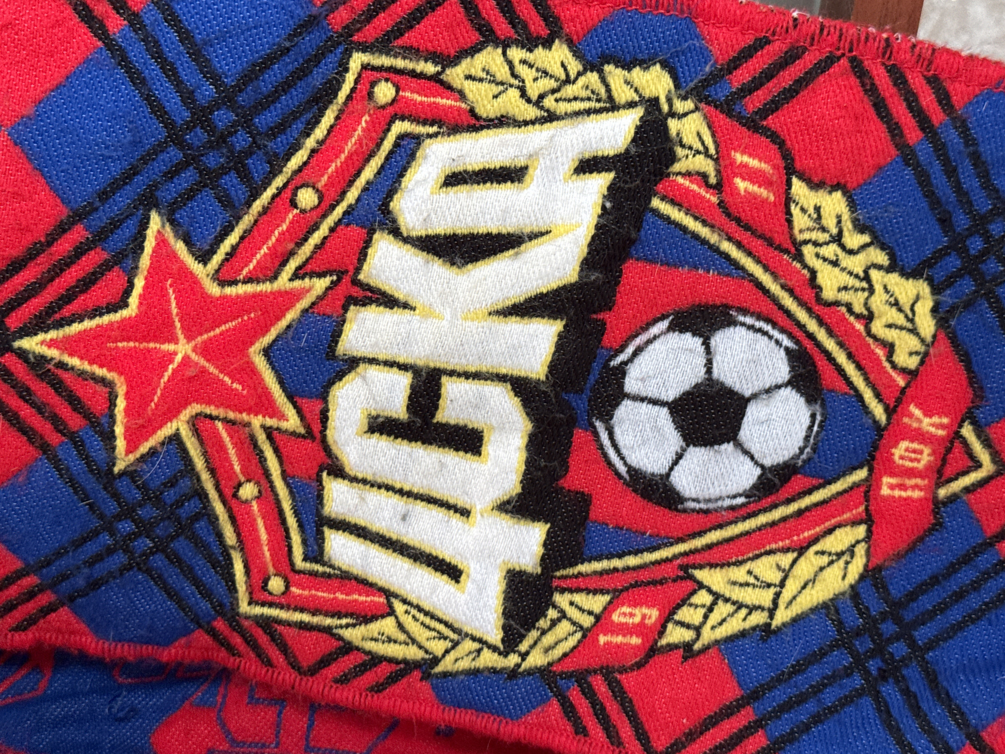 The image shows a close-up of a fabric patch or emblem, predominantly red and blue with a plaid pattern in the background. A central embroidered design is bordered by a red, diamond-shaped frame decorated with yellow leaves and what appear to be small dots. Within the frame, there is a five-pointed red star on the left and the letters "ИЖКА" in white with black outlining in the center. To the right of the letters is a soccer ball. On either side of the soccer ball and below the star, is the date 1968 and Ф.К. written in Russian. The overall impression is of a soccer-related emblem or logo. I am unable to identify the location in Izhevsk.