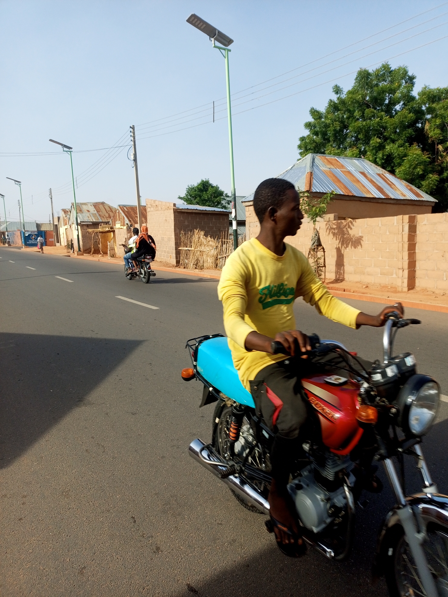 The image depicts a man riding a motorcycle on a paved road in the city of Gombe, Nigeria. He wears a long-sleeved yellow shirt and black pants, with his head turned to the left. The motorcycle has a red gas tank and a light blue seat. Another motorcycle carrying two people is further down the road. Buildings line the roadside, with corrugated metal roofs and walls constructed of what appears to be brick or mud. Streetlights are placed along the road. The sky is clear and blue, indicating daytime.