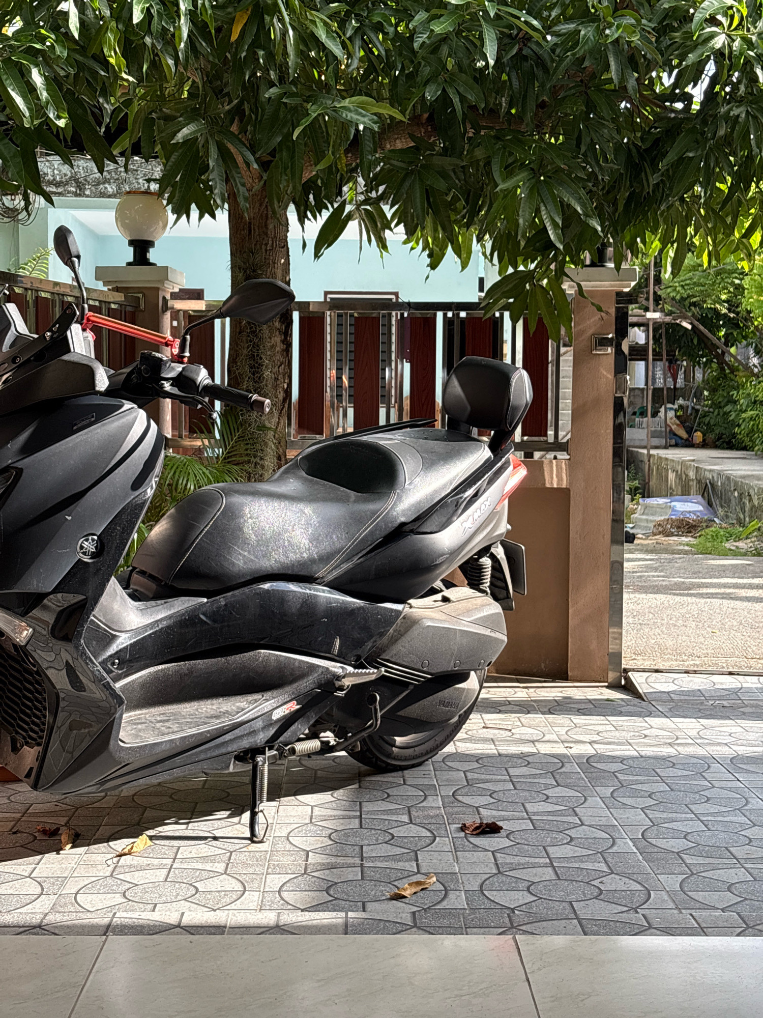 A black Yamaha XMAX scooter is parked on a tiled patio in front of a residential building, possibly in Choeng Thale, Thailand. The scooter is positioned at an angle, with its front to the left and rear to the right. It has a black leather seat and a backrest. Sunlight casts strong shadows from the scooter and a large tree with green leaves onto the patterned tiles. In the background, there is a fence and a glimpse of a street. The area appears to be outdoors, with tropical foliage visible.
