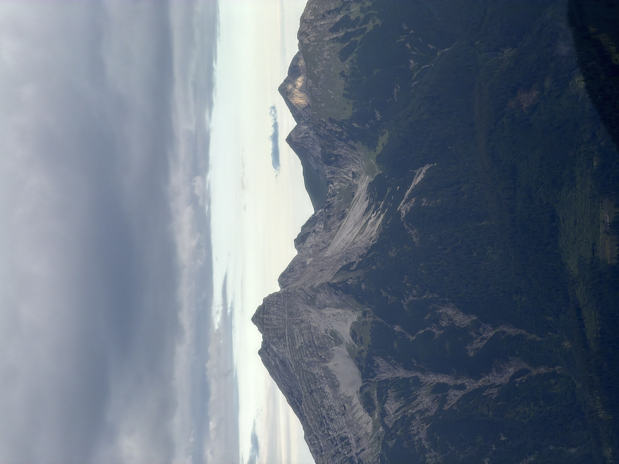 The image shows a majestic mountain range under a cloudy sky, likely in the vicinity of Grainau, Germany. The foreground is dominated by dark green coniferous forests that cover the lower slopes of the mountains. Above the treeline, the mountains transition to rugged, gray rock faces with visible geological strata and scree slopes. The peaks are sharp and jagged, indicating a high-altitude environment. In the distance, two prominent peaks rise sharply, with sunlight catching their upper slopes, contrasting with the shadowed valleys below. The sky is filled with a mix of dark, heavy clouds and lighter, more dispersed cloud formations, suggesting an overcast or impending weather change. No people or man-made structures are directly visible in the frame, emphasizing the natural grandeur of the landscape.