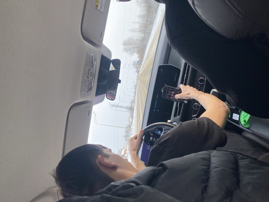 A person is driving a car on a snowy road, holding a phone in their right hand. The phone is mounted on the dashboard. The driver is wearing a black shirt and a puffy black jacket. The car's interior is visible, showing the dashboard, steering wheel, and rearview mirror. The exterior view through the windshield reveals a snow-covered landscape with bare trees and streetlights.