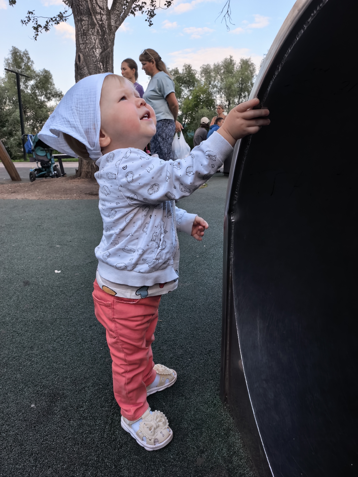 A toddler wearing a white headscarf, a light blue jacket with a cartoon pattern, and pink pants stands on a dark green playground surface. The child is looking upwards and to the right, with their right hand reaching out towards a large, dark, curved object, possibly part of a playground slide. In the background, several adults are visible. A tall tree with textured bark stands to the left of the child, and behind the tree, there are other trees and foliage. The sky above is a light blue with some wispy clouds, suggesting it is daytime. The overall scene appears to be a public playground. The image is taken from a low angle, focusing on the child and the playground equipment.