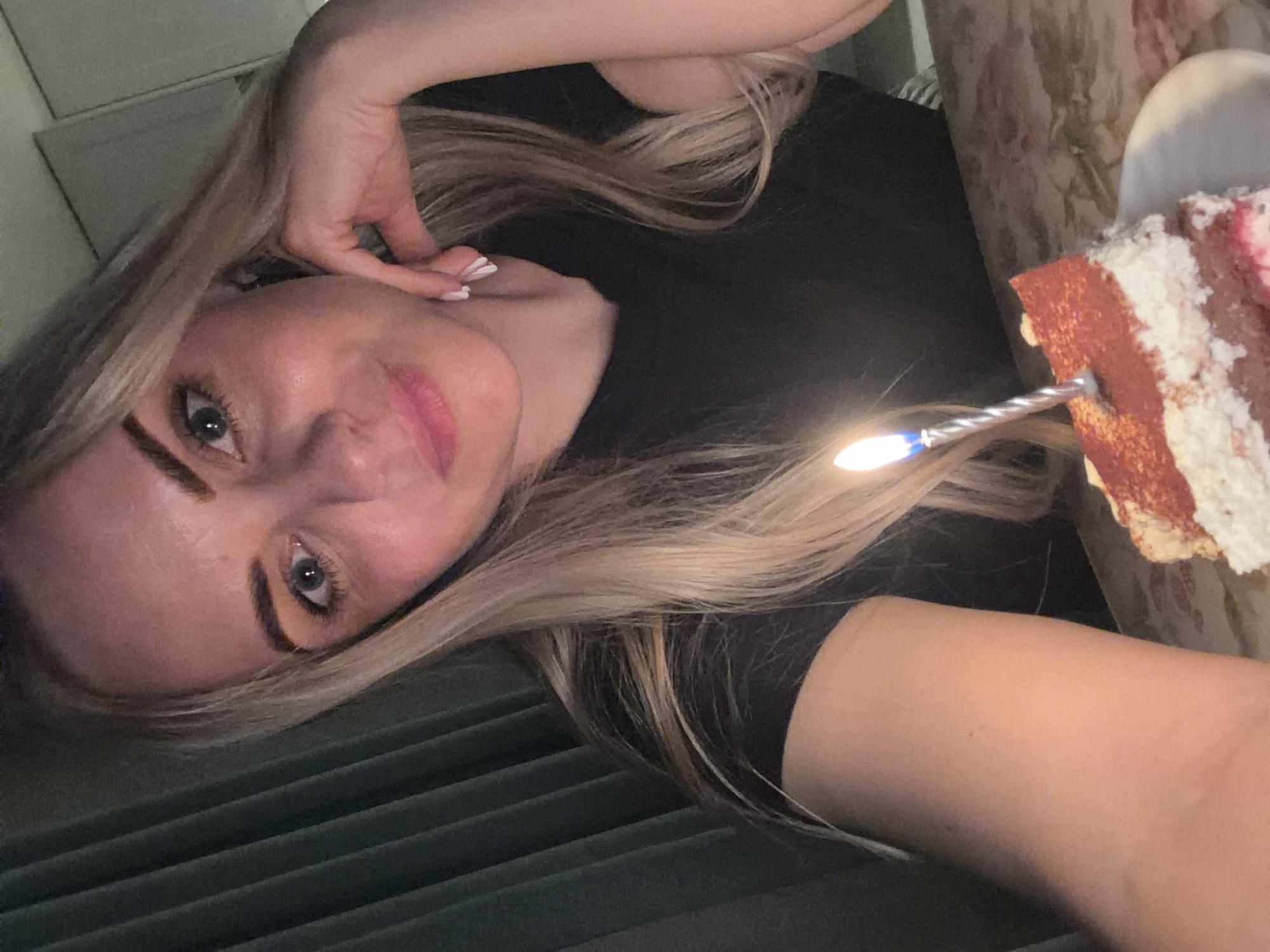 A young woman with fair skin, blue eyes, and blonde hair is holding a piece of cake with a lit candle in front of her. The cake appears to be red velvet with white frosting. She is wearing a black shirt. The background is indistinct, with a hint of blinds and a floral-patterned surface. The photograph appears to be taken indoors. The information provided is insufficient to determine the location in the city of Votkinsk, Russia.