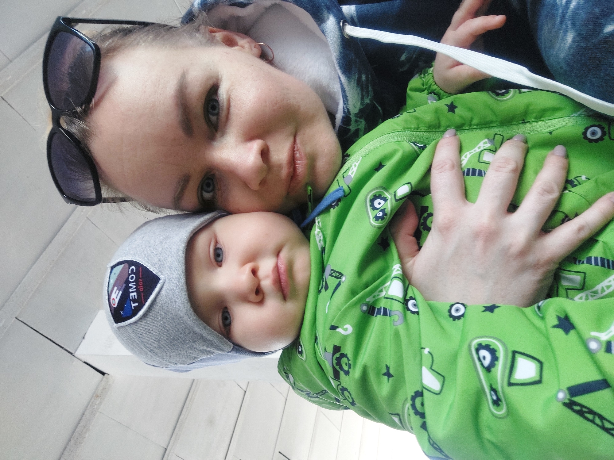 A young woman and a baby are posing for a photo. The woman has sunglasses on her head and is wearing a hooded sweatshirt. The baby is wearing a gray hat and a green patterned jacket. The background appears to be a white wall. The location is Chaikovsky, Russia.