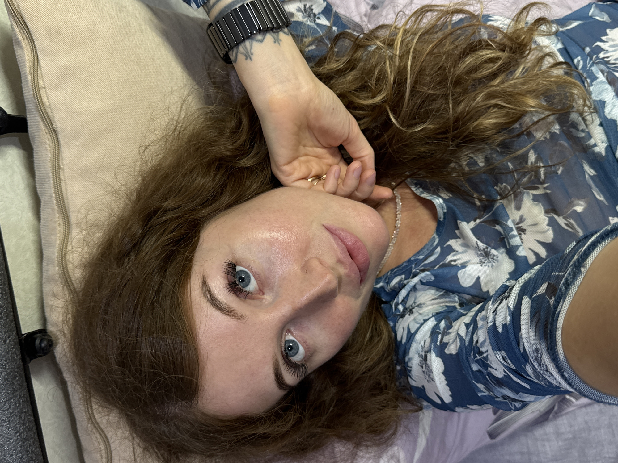 A light-skinned woman with brown hair and blue eyes is lying on her side on a light-colored pillow. She is wearing a blue floral top, a silver necklace, and a black watch. Her hand is near her face. The location is not evident in the image.
