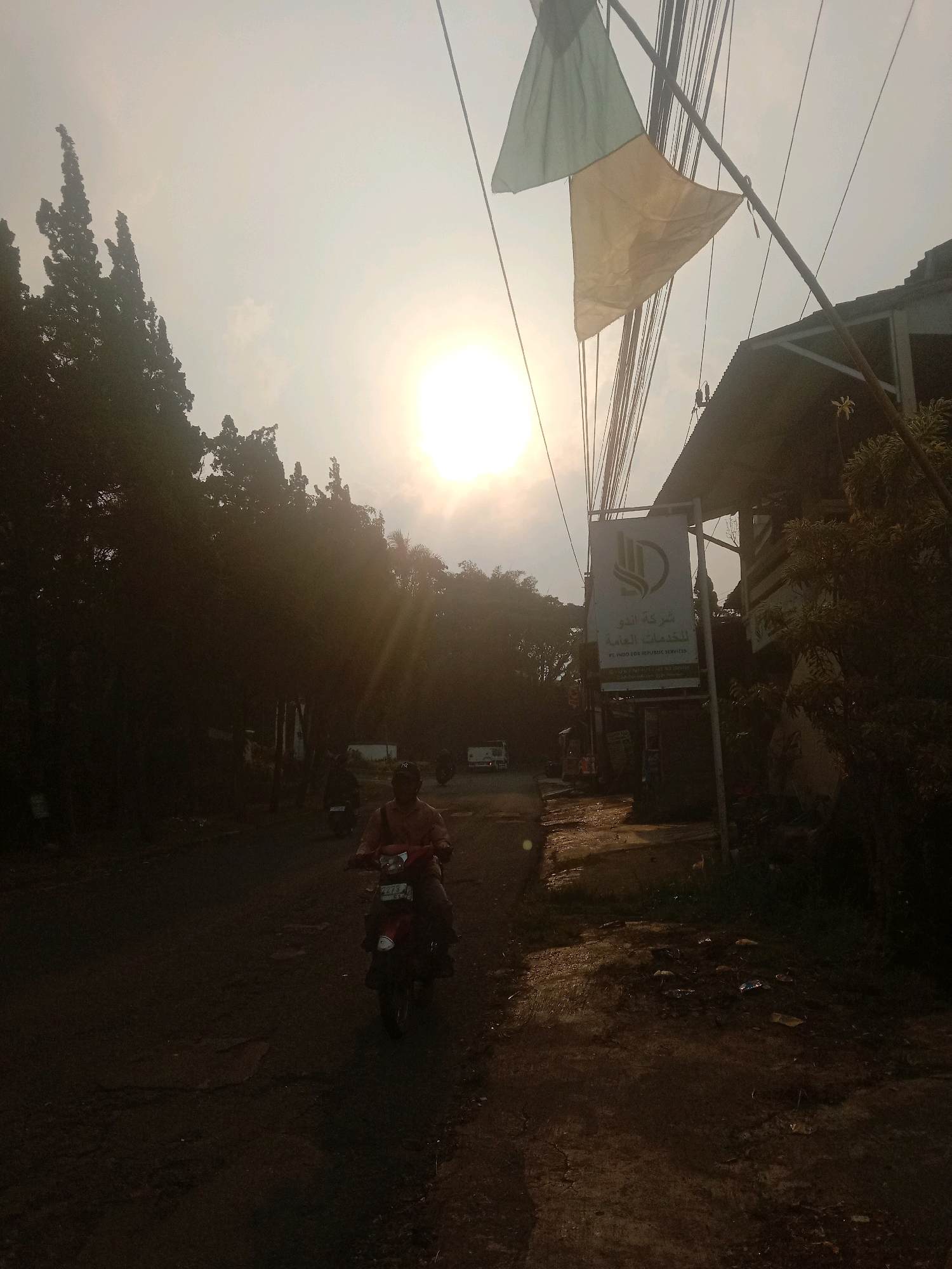 A person rides a red scooter on a dirt road in Palasari, Indonesia, during the day. The sun is high in the sky and partially obscured by clouds, casting long shadows. Trees line the left side of the road, and buildings are visible on the right. A sign in Arabic and English is mounted on a pole on the right side of the road. The road is unpaved and appears to be in disrepair. There are other vehicles and people visible in the background, suggesting a populated area. The overall atmosphere is hazy and warm, typical of a tropical climate.