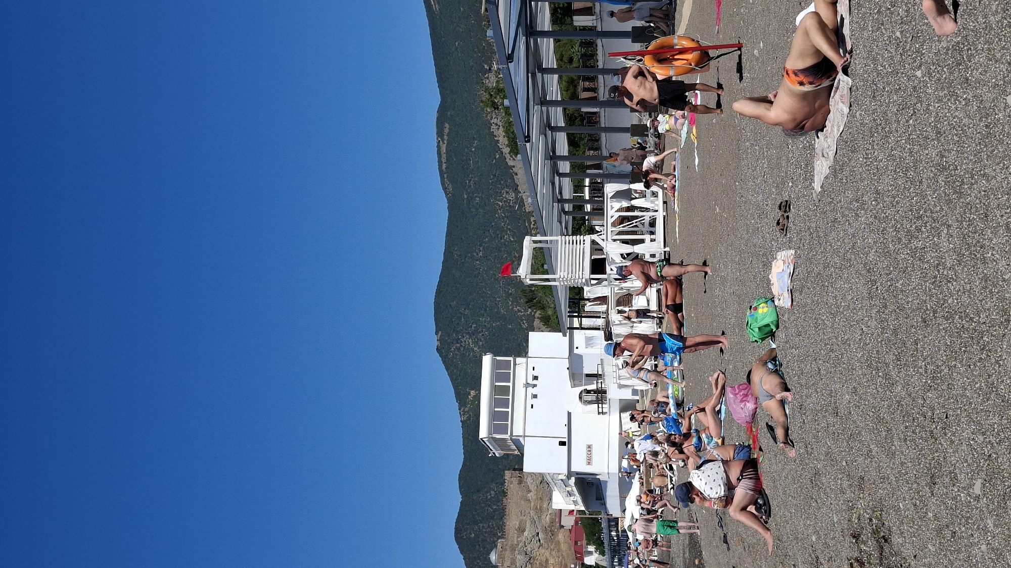 The image depicts a crowded beach scene under a clear blue sky. In the foreground, several individuals are relaxing on the pebble beach, some sunbathing, others sitting. They are dressed in swimwear. Various beach accessories such as towels, bags, and sandals are scattered around.
In the midground, there is a white building with multiple windows and signage that appears to read "MACCA". Adjacent to this building is a white lifeguard tower with a red flag flying at its top. Further back, there are covered seating areas and more people.
The background features a rocky hillside with a large, ancient-looking stone structure or fortress on the left. Beyond that, rolling green hills rise against the horizon. The sea is not visible in this frame. The overall atmosphere suggests a busy, sunny day at a popular beach resort.