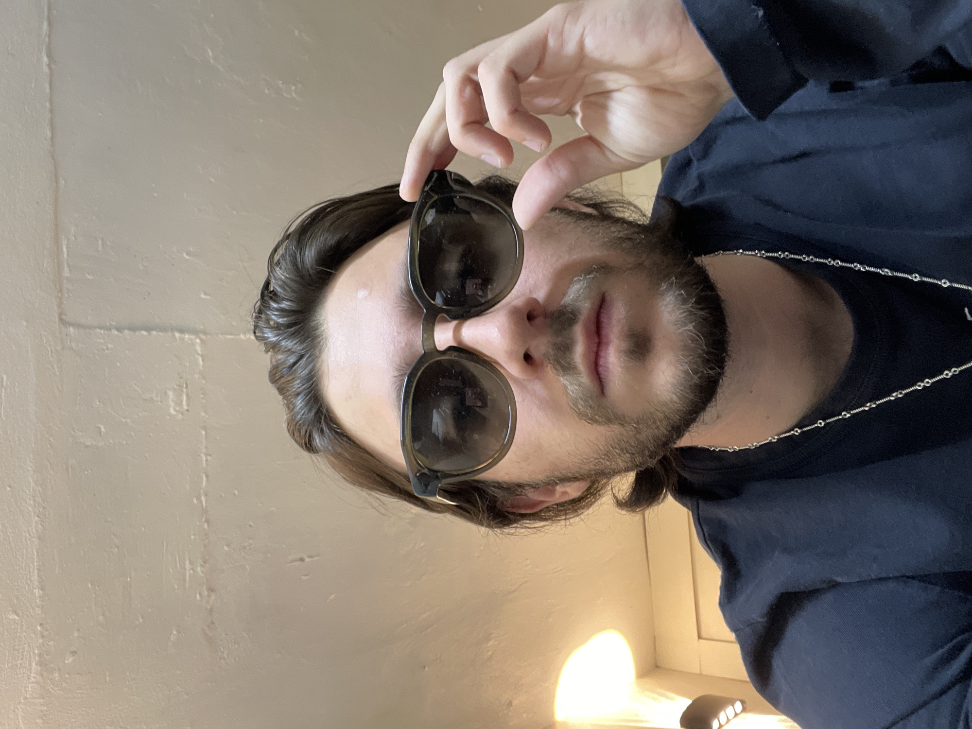 A medium shot depicts a man with long, dark hair and a beard, wearing dark sunglasses. He is holding the sunglasses with his right hand. He is wearing a dark-colored, long-sleeved shirt and a silver chain necklace. The background is a beige wall. The location is unknown and cannot be determined from the image. The image does not contain any information that can tie it to Huixquilucan de Degollado, Mexico.