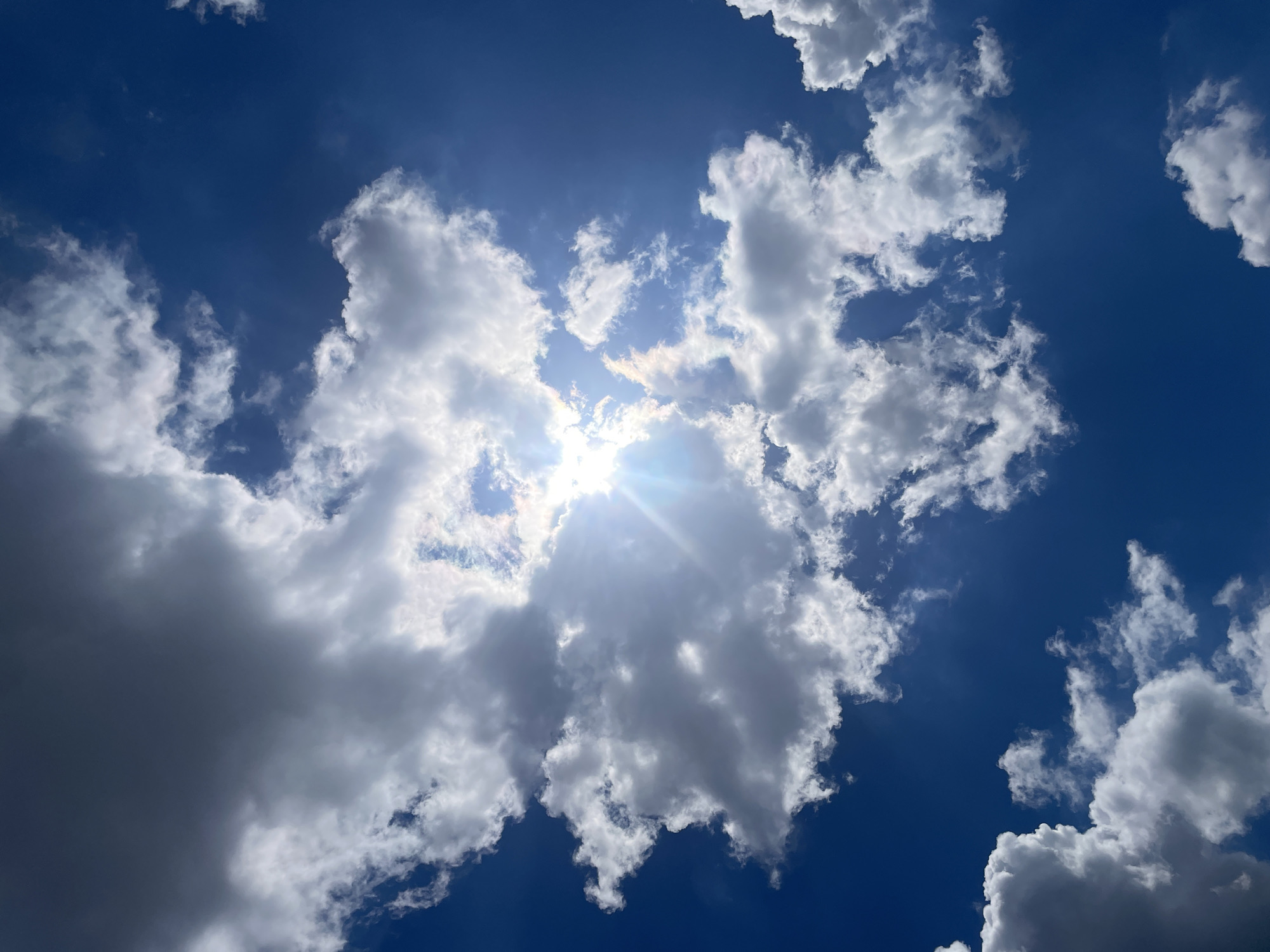 The image is a close-up, low-angle shot of a bright blue sky filled with scattered white and gray clouds. The sun is prominently visible, partially obscured by a large, puffy cloud, and casting bright rays and lens flare into the frame. The clouds vary in density, with some appearing wispy and translucent, while others are thick and shadowed. The overall impression is one of a sunny, partly cloudy day. There are no people, animals, buildings, or any other objects visible in the frame, making the scene abstract and focused solely on the sky and clouds. The location is stated as Unknown, Unknown.
