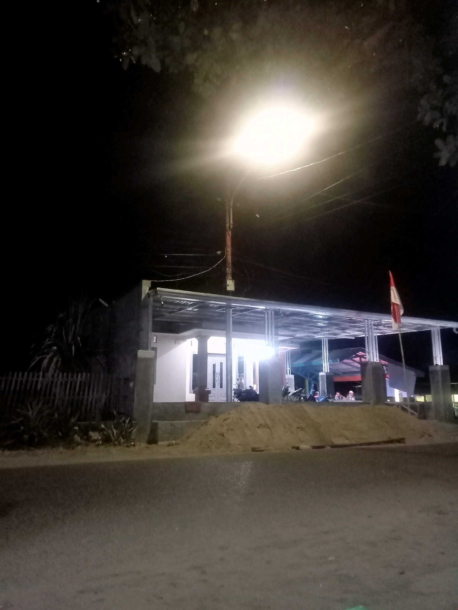 This is a night-time photograph taken from a street, likely in Mongolato, Indonesia, looking towards a partially constructed building. A bright streetlamp illuminates the scene from the top left, casting harsh light and creating strong shadows. In the foreground, a paved road is visible, with some scattered debris and a faint green object on the right side.
The building appears to be a single-story structure with a modern design, featuring a metal roof supported by pillars. The front of the building has large white doors and windows, and the interior is brightly lit, revealing some furniture and what appear to be ceiling fans. To the left of the building, there is a wooden fence and dense foliage. In front of the building, a pile of sand is visible, suggesting ongoing construction.
On the right side of the building's facade, an Indonesian flag is mounted on a pole. Several motorcycles are parked under the covered area of the building, and a few individuals are seated at tables inside, illuminated by the building's interior lights. The background is dark, typical of a night sky, with some electrical wires running across the frame, presumably connected to the streetlamp. The overall impression is of a scene in a developing area at night, with a mixture of residential and possibly commercial activity.