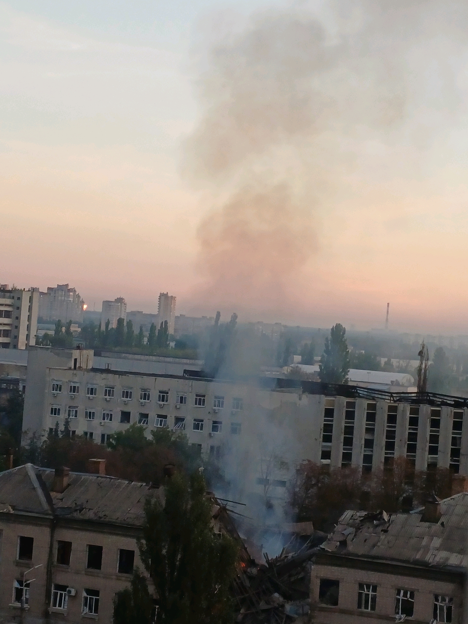 The image depicts a cityscape with smoke rising into the sky, suggesting a fire or explosion. In the foreground, there are several buildings, some of which appear to be damaged, with debris visible on rooftops. The buildings are multi-story structures with numerous windows. Trees are interspersed throughout the scene, and in the background, taller residential buildings and industrial structures are visible. The sky is overcast with a soft, diffused light, indicative of either early morning or late evening. The overall atmosphere suggests a scene of destruction or aftermath.