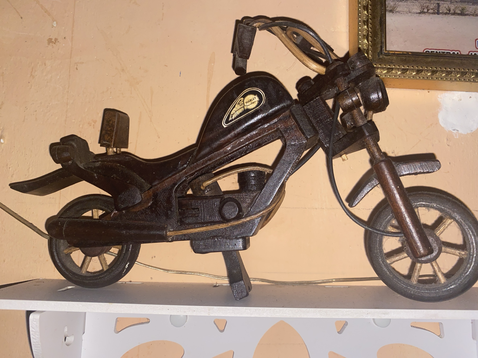 This is a side-view, medium shot of a wooden model of a motorcycle. The motorcycle is dark brown and appears to be handmade. It has a detailed design, including a visible engine, handlebars, wheels with spokes, a seat, and a kickstand. The motorcycle is positioned on a white shelf, and behind it is a peach-colored wall with a framed picture. The picture frame is ornate and gold-colored, and the image within it is unclear but appears to be a landscape. A portion of a second, less ornate frame is visible to the left of the gold frame. The lighting suggests it is daytime, indoors. There is no discernible weather or emotional context. A small label with a white background and a black outline is visible on the side of the motorcycle's fuel tank, featuring an image resembling a shield with wings and the text "HARLEY DAVIDSON" inside. The overall scene is a static display of a decorative object.