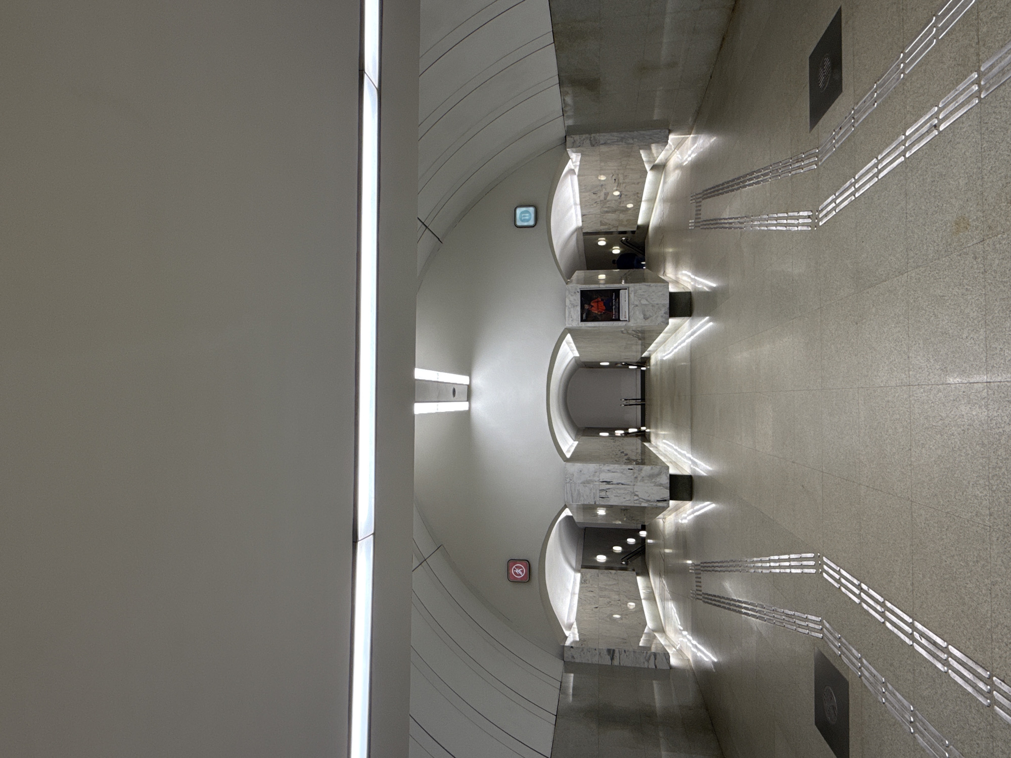 The image depicts an interior shot of a Moscow Metro station in Russia. The architecture features a clean, modern design with curved walls and a bright, neutral color palette. The station has multiple levels connected by staircases. Textured flooring and tactile paving is installed to assist visually impaired passengers. Signage is present, including a "no smoking" symbol and metro line signs. The lighting is primarily artificial, illuminating the walls and floor. There are no visible people in the scene.