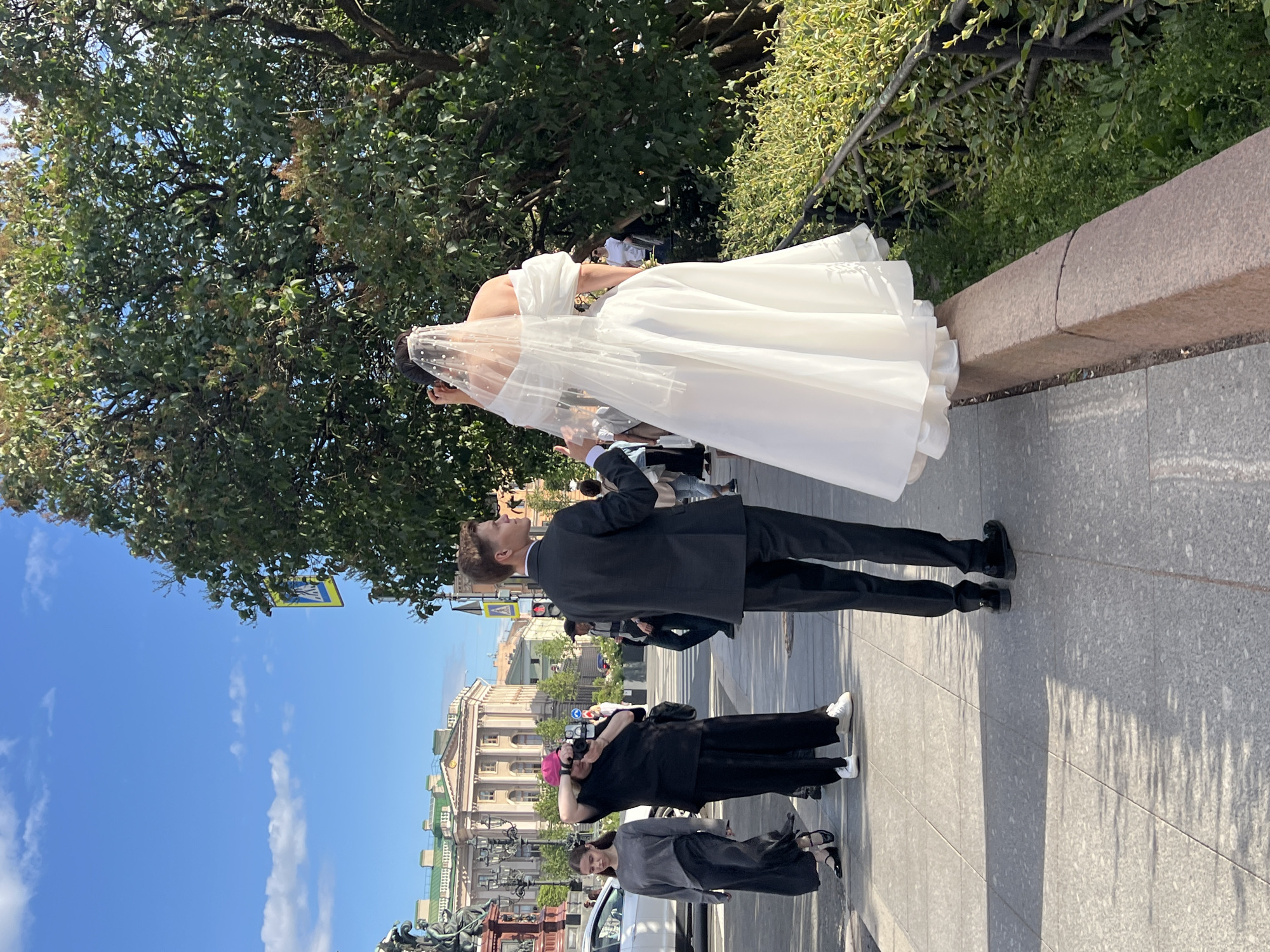 In Saint Petersburg, Russia, on a sunny day, a bride in a white off-the-shoulder wedding dress and veil stands on a stone barrier, facing a tree. Beside her, a man in a dark suit faces away from the camera. In the background, on the sidewalk, a woman in a grey dress walks past a person wearing a pink hat and black clothing who is taking a photograph with a camera. A white car is parked on the street. Behind them, a building with classical architecture and green accents is visible under a blue sky with scattered white clouds. The scene is set along a paved sidewalk lined with a low metal fence and lush green bushes.