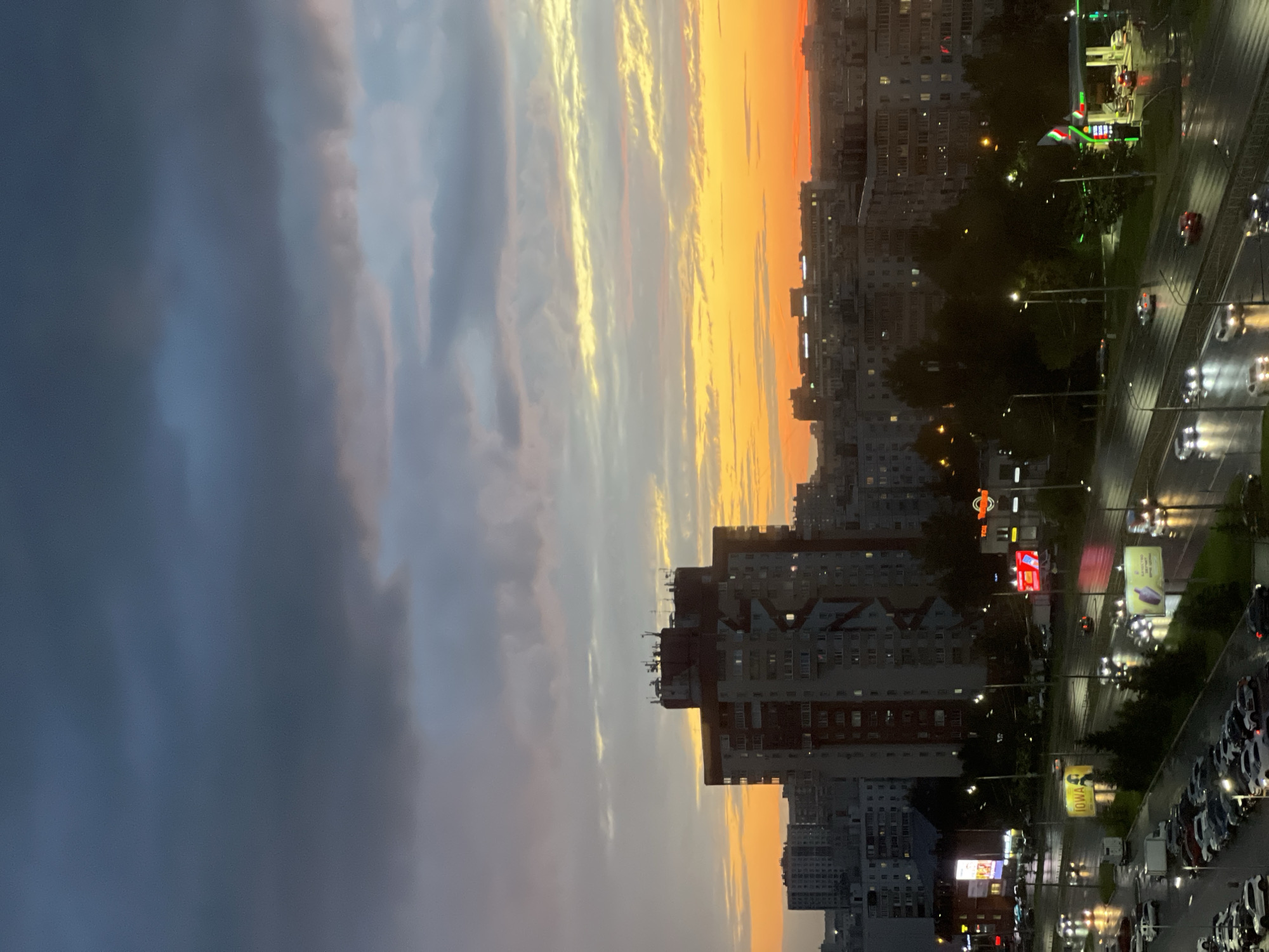 The image depicts a twilight aerial view of an urban environment, likely Kazan, Russia. Dark, heavy clouds dominate the upper portion of the sky, with a vibrant band of orange and yellow light from the setting sun breaking through near the horizon. Below the sky, a cityscape unfolds, characterized by a cluster of mid-rise and high-rise apartment buildings, one particularly prominent, with many windows illuminated, suggesting evening or night.
A multi-lane road is visible in the foreground, wet from recent rain, with the reflections of streetlights and vehicle headlights shimmering on its surface. Several cars are in motion on the road, their headlights and taillights creating streaks of light. On the right side of the road, a brightly lit gas station with a green and red canopy is operational, with cars visible at the pumps. Billboards and commercial signage are present along the roadside, some illuminated. The ground level features parked cars in a lot to the left. Trees and vegetation are interspersed throughout the urban landscape, particularly along the road and around buildings. The overall atmosphere is one of an overcast evening in a bustling city.