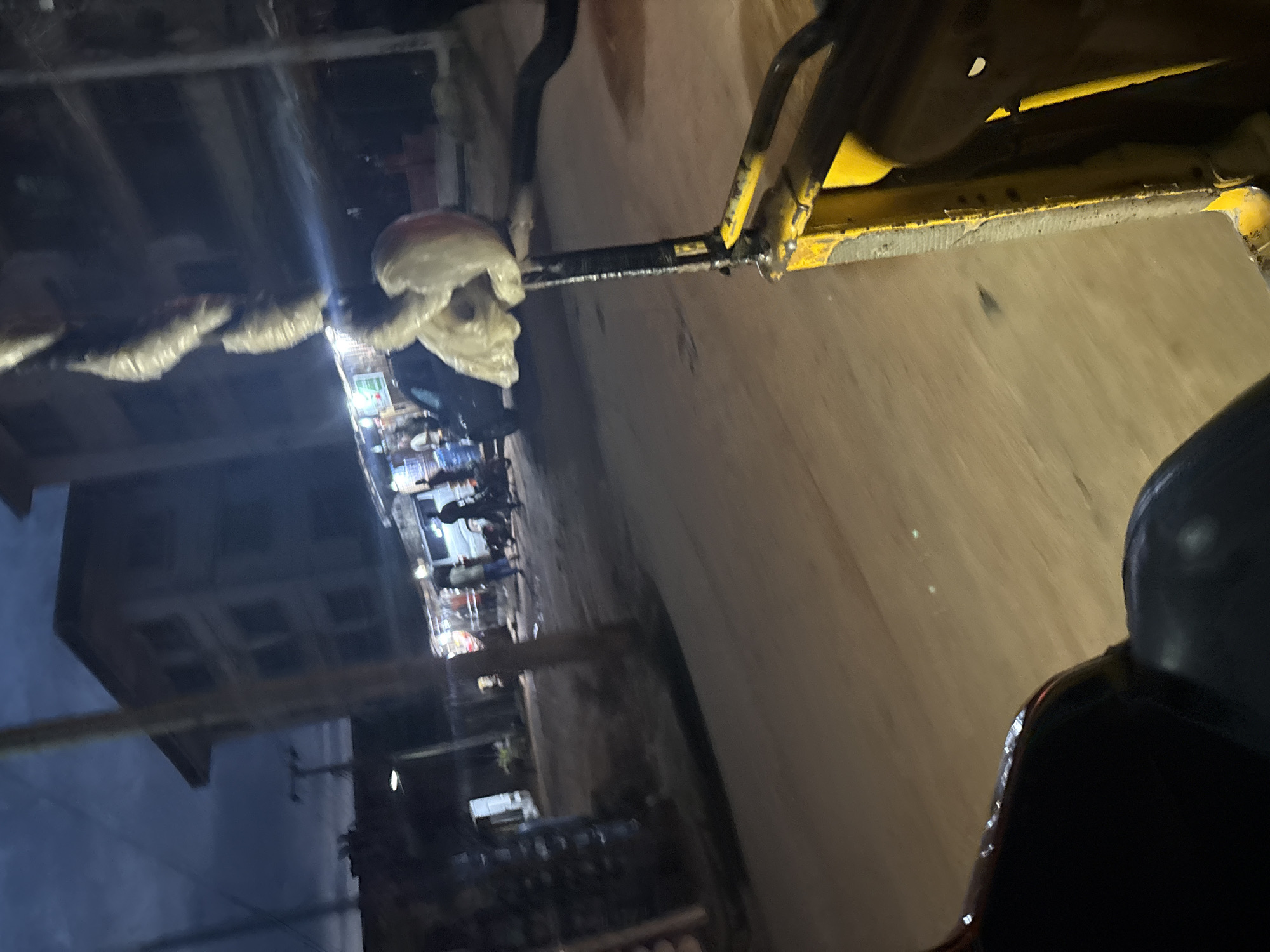 The image depicts a street scene at dusk or night in Onitsha, Nigeria. In the foreground, a portion of a yellow vehicle, possibly a rickshaw or tricycle, is visible, suggesting the image was taken from within or behind this vehicle. A dark, plush seat is also in the foreground on the left. A vertical metal pole with wrapped objects, possibly goods in plastic, attached to it extends upwards from the vehicle.
The background reveals a street lined with buildings and market stalls. Several individuals are present, some standing and others appearing to be in motion. A dark-colored car is parked on the side of the road. Streetlights and lights from the market stalls illuminate the scene. The sky is dark, indicating nighttime or heavy cloud cover. The overall atmosphere is that of a bustling urban environment at night.
