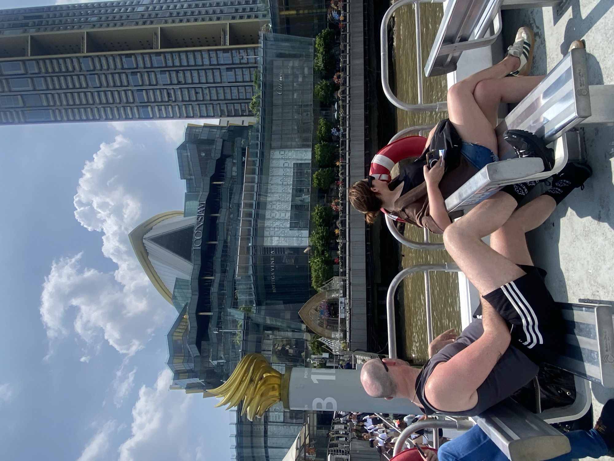 The image shows a scene in Bangkok, Thailand, taken from a boat on the Chao Phraya River, featuring the iconic ICONSIAM shopping mall in the background. The foreground is dominated by two people relaxing on the boat seats. One person wears black Adidas shorts and a gray tank top, while the other wears denim shorts, a brown shirt, and has a red life preserver nearby. The river water is a murky brown. Several other boats and people can be seen along the riverbank. The sky is bright with fluffy white clouds. A golden torch structure stands near the dock.