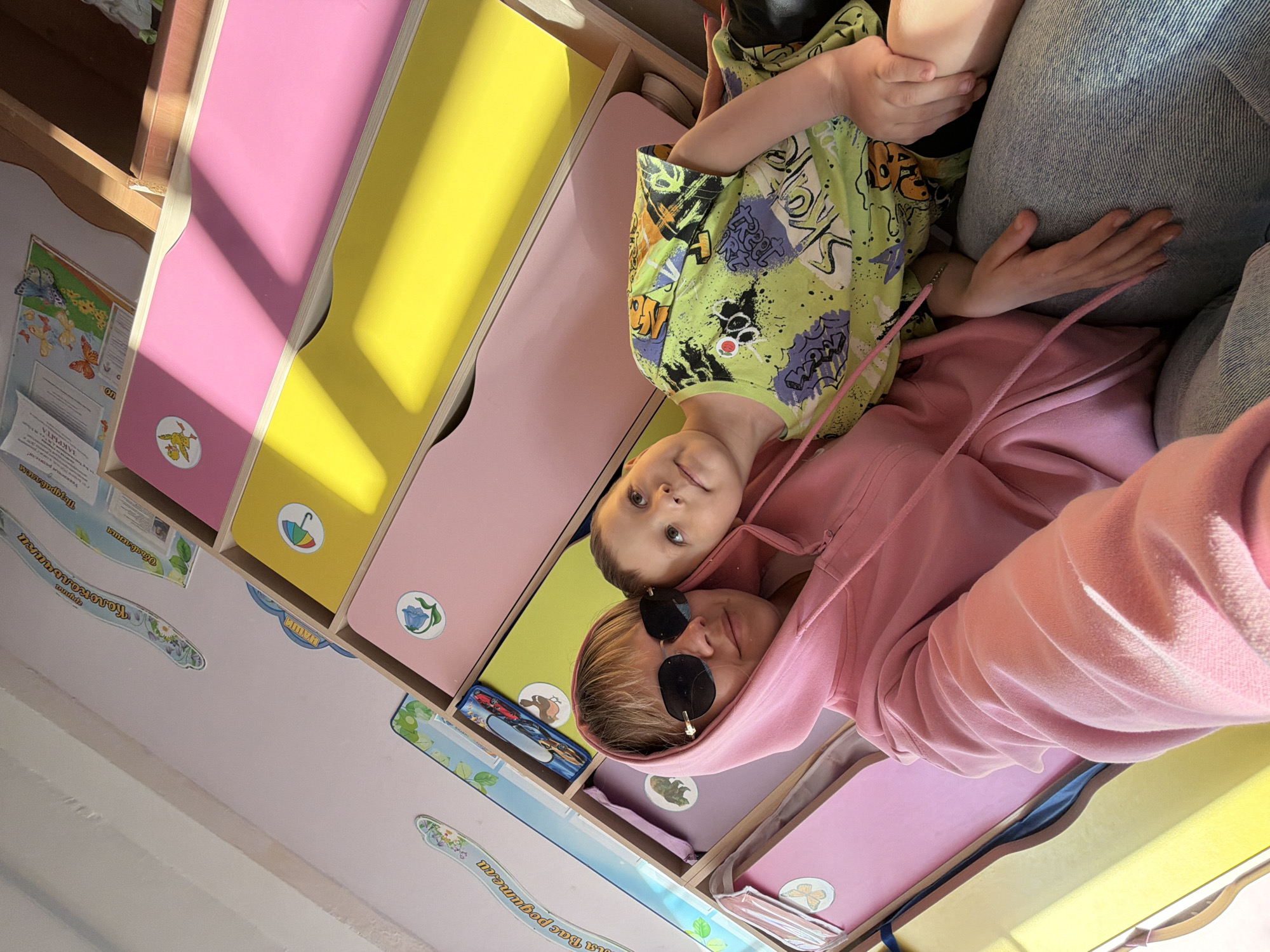 Captured indoors, the image presents a scene featuring a woman in a pink hooded sweatshirt and sunglasses, alongside a young child in a green patterned shirt and jeans. They are situated next to a wall-mounted set of colorful storage drawers, with pink and yellow being the predominant colors.
The location of this image is identified in Chaikovsky, Russia, based on the presence of a sign partially visible at the lower left edge of the image, which appears to contain Russian text, specifically the phrase "для Вас родители" (for you, parents). This type of text is typical of informational signage in public spaces or institutions like schools or kindergartens in Russia. The phrase "ЗАКРЫТА" (Closed) is also visible on one of the other signs.
Details on the wall surrounding the storage drawers include decorative stickers and informational signs, indicative of a child-friendly environment, such as a nursery or playroom. The woman and child are positioned close together, suggesting a familial relationship.
The photograph is taken from a slightly tilted perspective, potentially captured by the woman herself, indicating a spontaneous moment within an indoor setting in Chaikovsky, Russia.