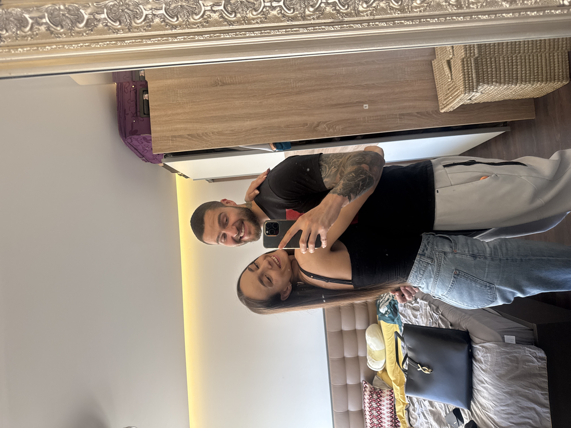 A mirror reflects a couple taking a selfie indoors. The man has a beard and is wearing a black t-shirt and grey sweatpants. His arms are covered in tattoos. The woman has long brown hair, wears a black top, and blue jeans. They are smiling at the camera held by the man. The reflection reveals a room with a wooden wardrobe and a basket on top. A bed with various items, including a black bag and slippers, is visible. A light fixture illuminates the wall next to the bed. This scene takes place in Sofia, Bulgaria.