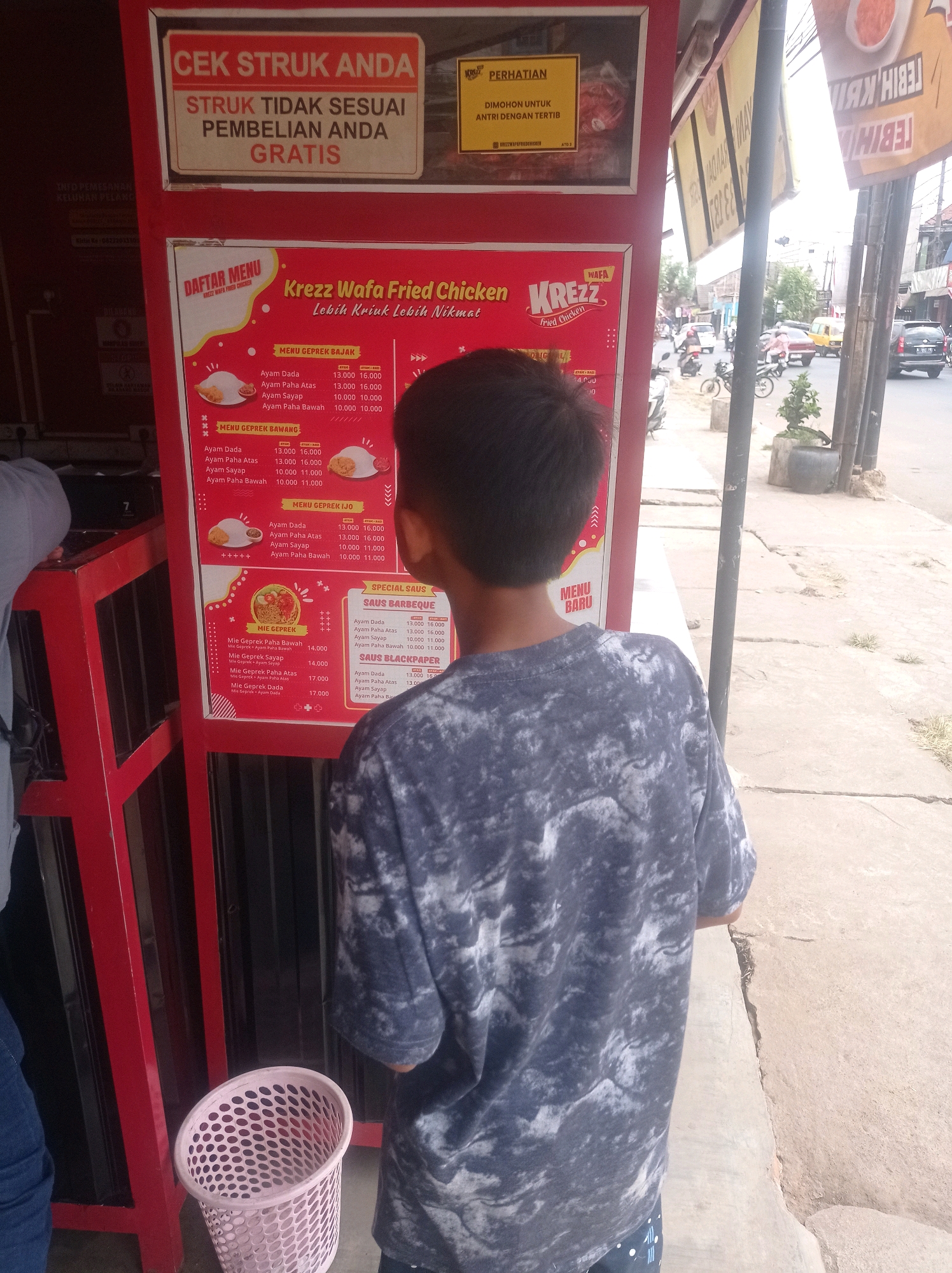 The image shows an outdoor view of a food stall in Bululawang, Indonesia. A young person, viewed from behind, is standing in front of a large red menu board. The menu board displays prices for various chicken dishes, including "Menu Geprek Bajak," "Menu Geprek Bawang," "Menu Geprek Ijo," and "Mie Geprek." There are also "Saos Barbeque" and "Saos Blackpaper" options. To the left of the menu board, a sign reads "CEK STRUK ANDA" and "STRUK TIDAK SESUAI PEMBELIAN ANDA GRATIS." Further to the left, a trash can is visible. In the background, a street scene with passing vehicles and buildings can be seen. The stall appears to be part of a small commercial area.
