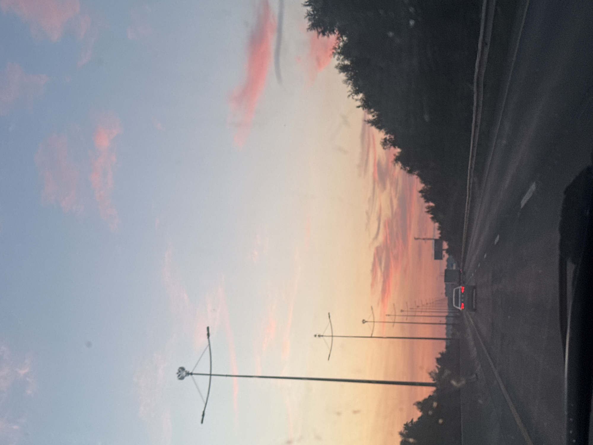 The image captures a sunset view from a car driving on a highway in Kondratovo, Russia. The sky is a gradient of light blue and pink, with streaks of pink clouds. A line of streetlights runs along the left side of the highway. On the right side, a dense line of dark green trees borders the road. A white car is visible in front of the viewer's vehicle, driving in the same direction. Further ahead, a truck can be seen. The highway has visible white lane markings. The overall scene depicts a serene, late-day journey.