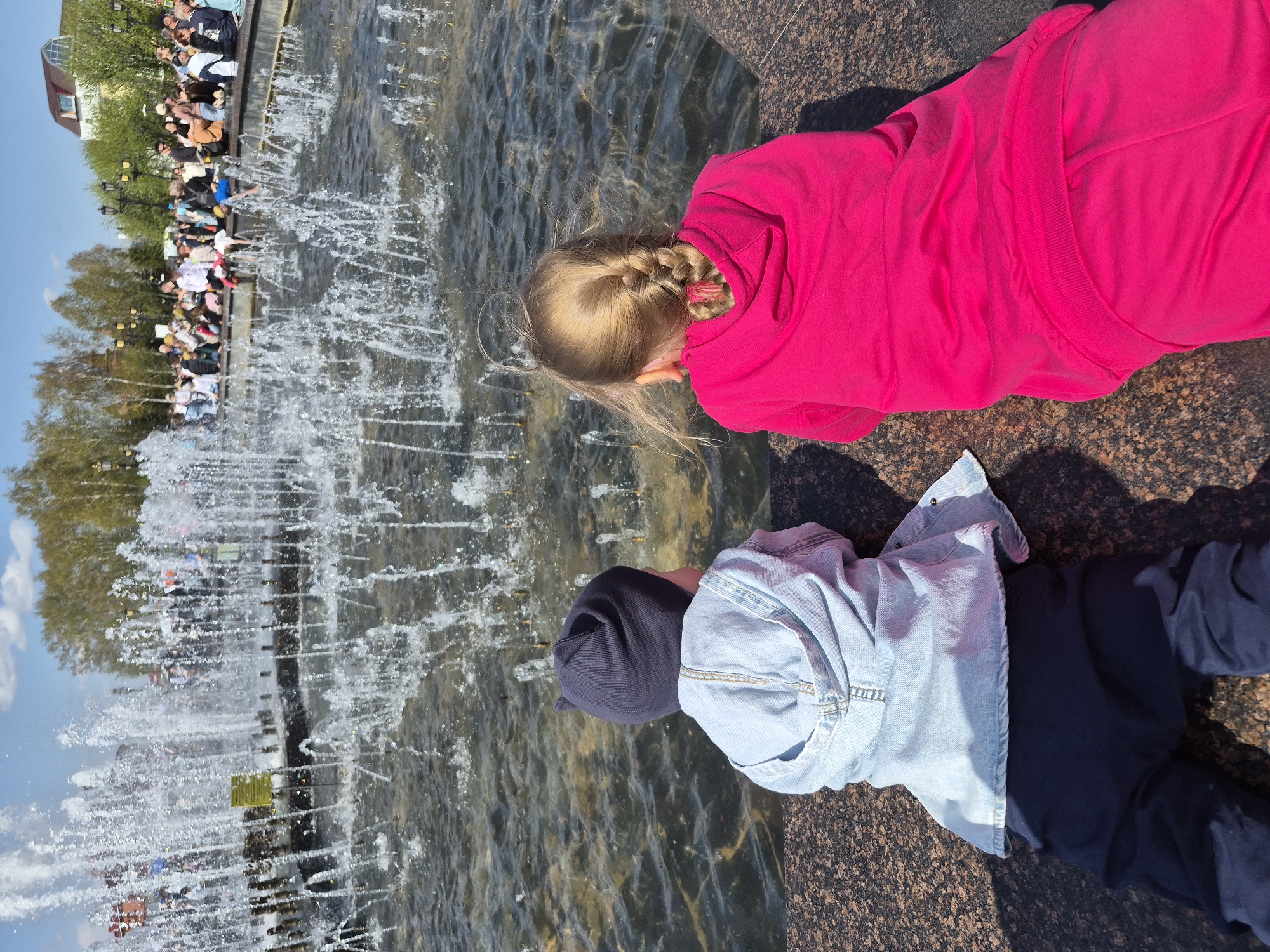 Two children are lying down near a fountain. One child wears a pink hoodie, has blonde hair with a braid, and faces the fountain. The second child wears a black beanie, a light blue denim jacket, and dark pants, also facing the fountain. The fountain has multiple water jets spraying upwards. Behind the fountain, a crowd of people stands. Trees and a building are visible in the background. The location is Tobolsk, Russia. The ground is a dark, speckled stone.