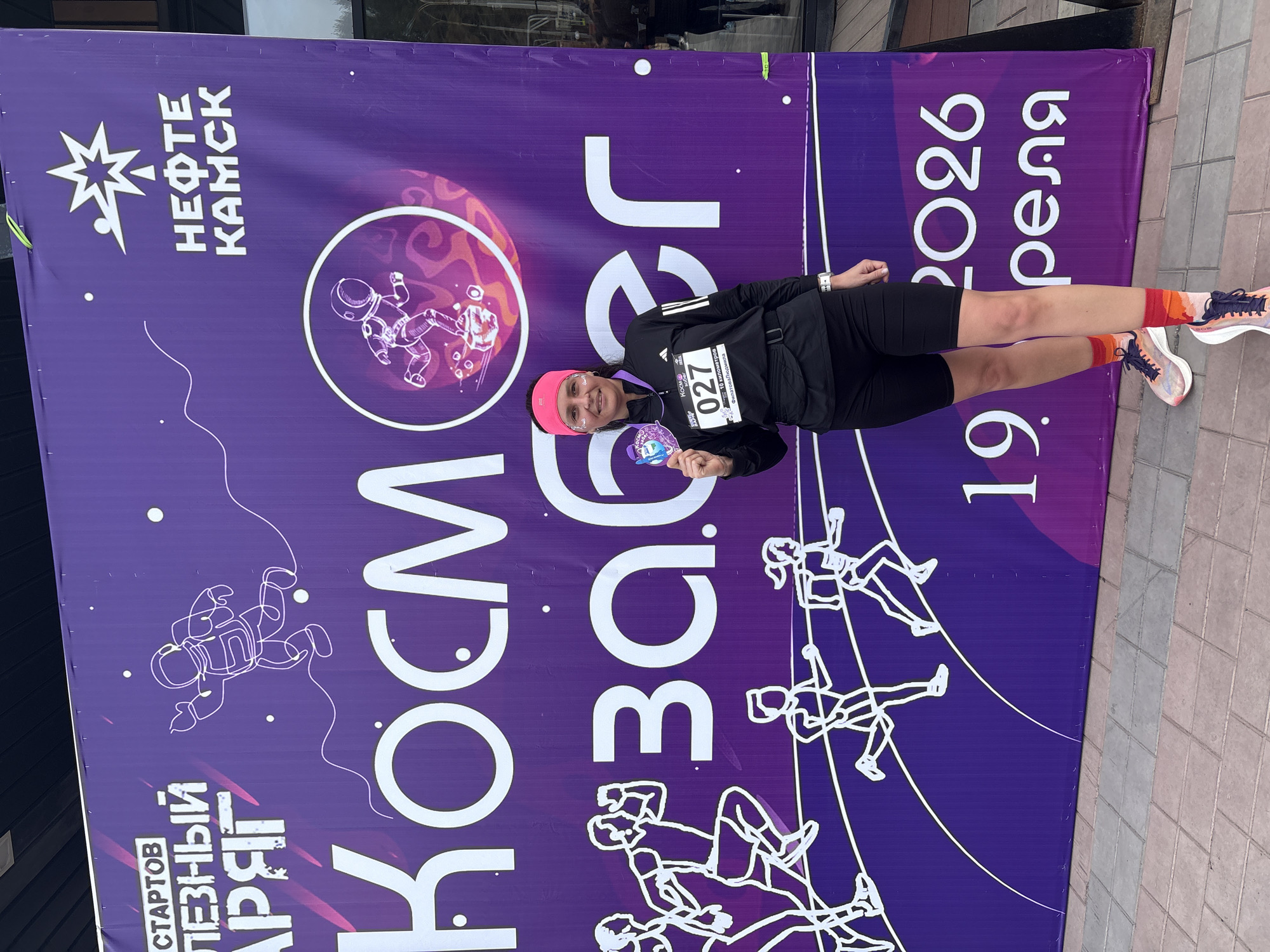 A woman stands smiling in front of a purple event banner. She is wearing a black Adidas jacket over athletic clothing and is holding up an event medal in her left hand. Her race number, 027, is pinned to her jacket. The banner is a backdrop for the "Cosmos Run" event, as indicated by the large Cyrillic text "Косм забег". The city "НЕФТЕ КАМСК" (Neftekamsk) is printed at the top left of the banner, identifying the event location in Russia. An illustration of an astronaut and people climbing ropes are also on the banner, consistent with the space and athletic themes of the event. The date "19. 2026 реля" is also printed on the banner. The background suggests an outdoor event on a paved area.