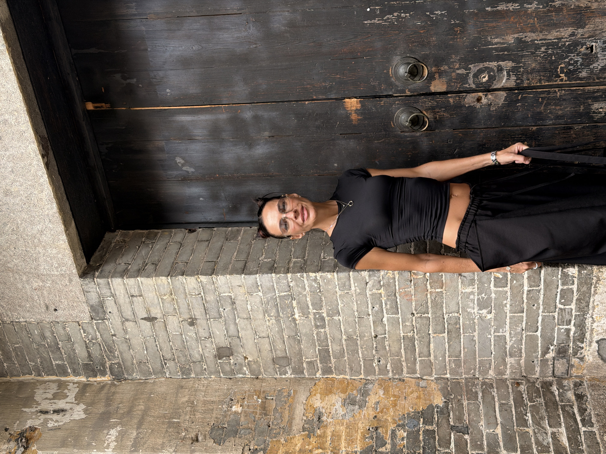 A woman stands in front of a large, weathered wooden door in Chancheng District, China. She is wearing a black crop top and black wide-leg pants, with her hair styled in two buns. She is also wearing glasses and a necklace. The door has two large, ornate brass handles and appears to be very old, with visible wear and tear. To the left of the door is a brick wall with peeling paint and patches of exposed plaster. The overall scene suggests an outdoor setting in an area with historical architecture.