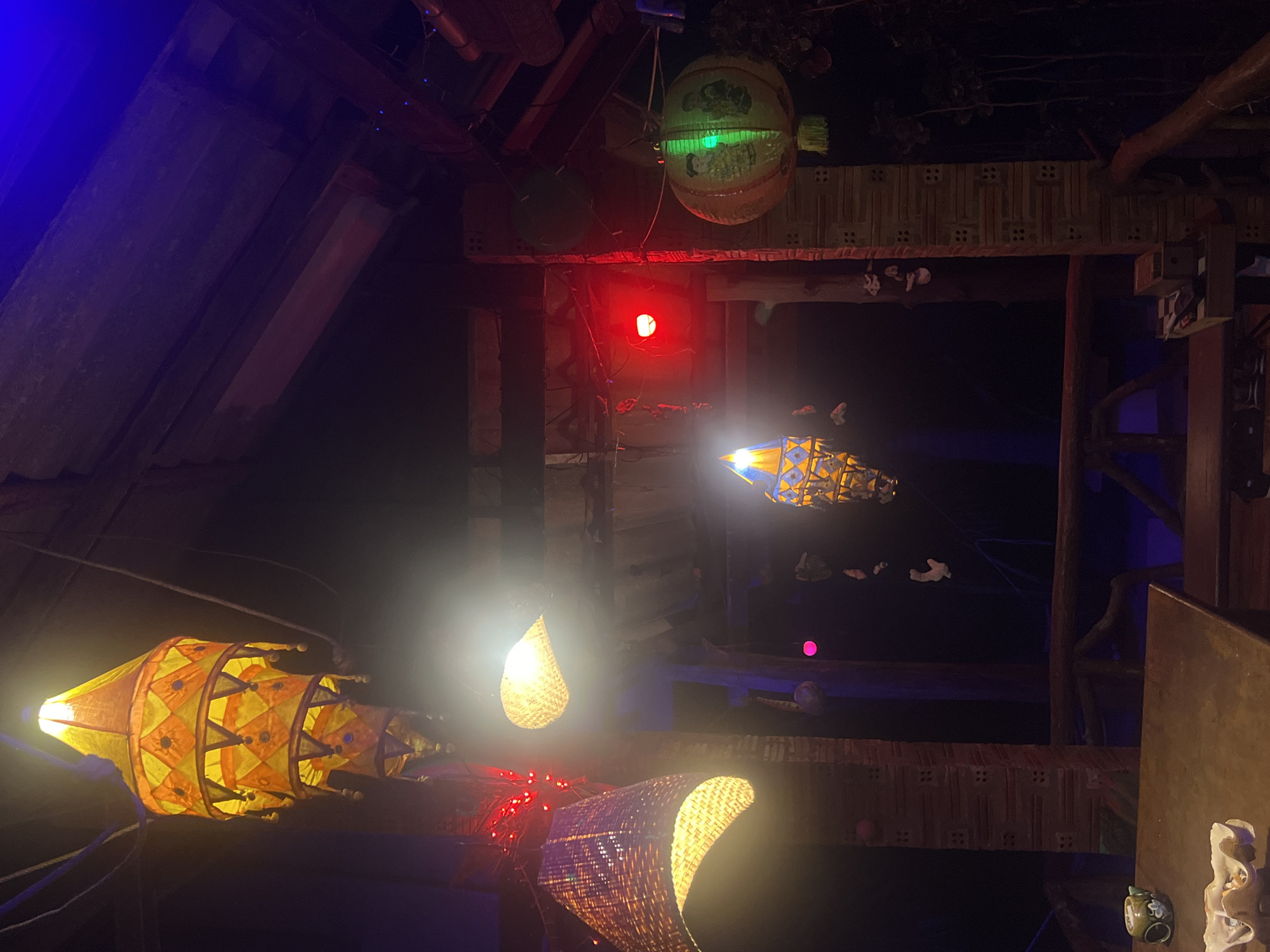 The image captures a dimly lit, indoor scene with various decorative lights and objects. The setting appears to be a rustic or bohemian-style establishment, possibly a bar, restaurant, or art space, with exposed wooden beams and textured walls.
Dominating the foreground is a large, conical lantern with an intricate pattern of yellow and orange fabric, illuminated from within, casting a warm glow. To its right, a woven basket-shaped lamp also emits a bright light. Further in the background, a spherical lantern with a green glow, featuring some sort of imagery, hangs from the ceiling. A small red light is visible in the mid-ground, adding a splash of color to the otherwise dark environment. In the far background, through an opening, a silhouette of what looks like a floating or suspended object with lights can be seen.
The overall ambiance is mysterious and inviting, suggesting an evening or nighttime setting. The presence of multiple decorative lights creates a festive and possibly whimsical atmosphere. No people are clearly visible, but the arrangement of objects suggests a place designed for gathering or entertainment. The image is taken from a low angle, looking upwards and into the space. No discernible text is present in the image.