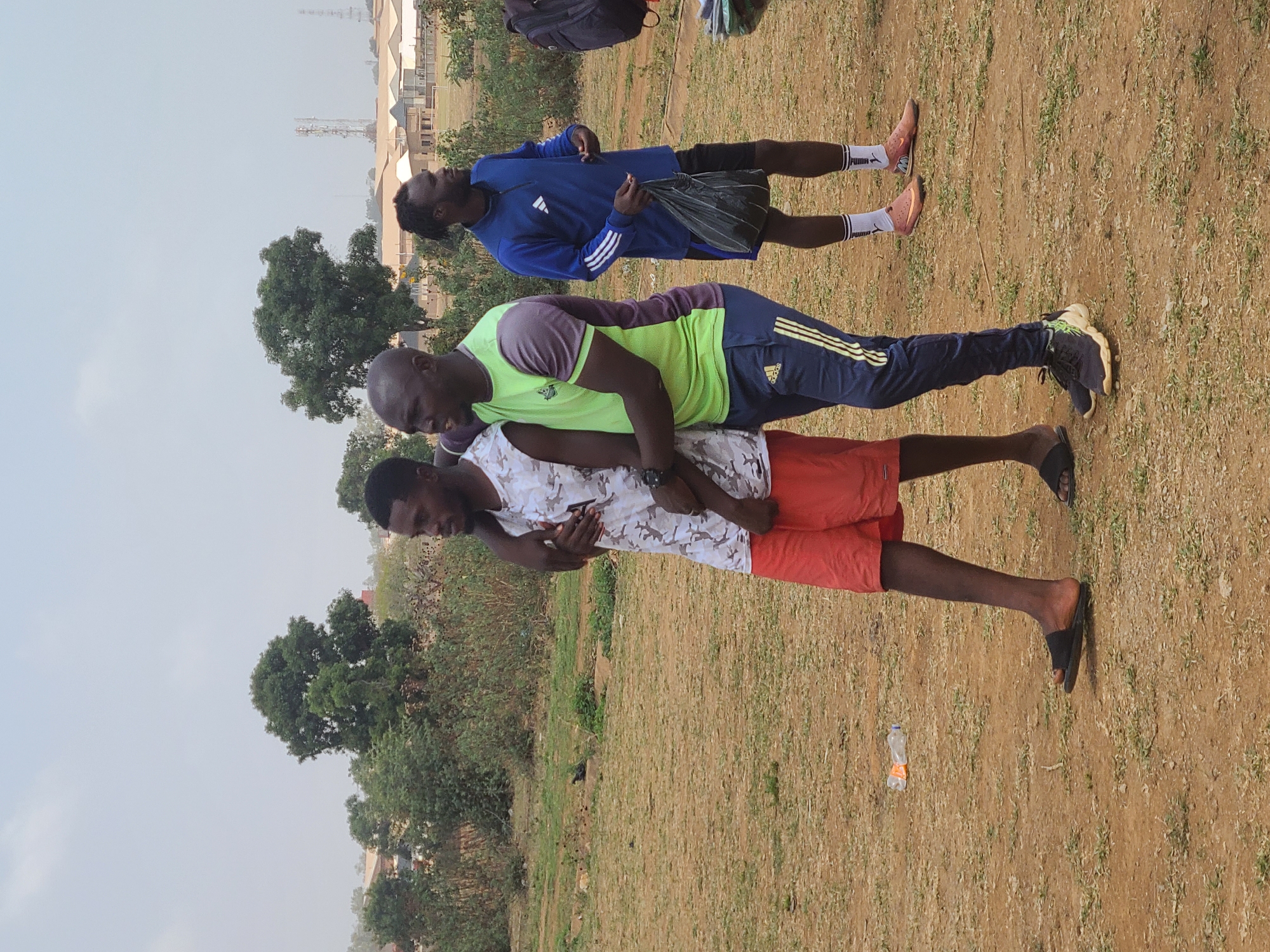 The image depicts three individuals in an outdoor setting, likely a field or grassy area, in Barikin Jirga Summa, Nigeria. The foreground shows a man in red shorts and a white patterned tank top supporting another man in blue Adidas pants and a light green t-shirt on his back. The second man is in a plank position, supported by the first man. A third man in a blue Adidas tracksuit is lying on the ground behind them. The ground is primarily dry grass and bare earth. There are some trees and bushes in the background, as well as buildings and structures further in the distance. A plastic bottle is visible near the feet of the man in red shorts.