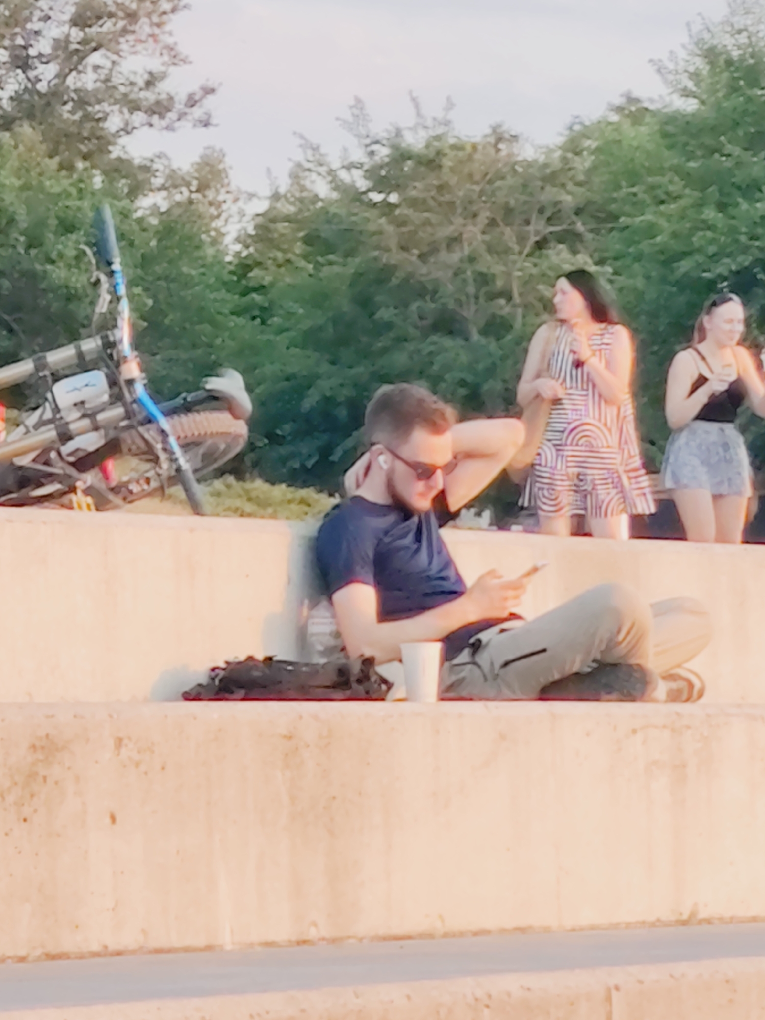 The image shows a man sitting on a concrete ledge, looking at his phone. He is wearing a dark blue t-shirt, light-colored pants, and dark shoes. He has earbuds in and sunglasses on his face. To his left, a white cup is visible, and to his right, a dark object is placed on the ledge. Behind him, two women are standing; one is wearing a striped dress, and the other is wearing a black top and a patterned skirt. In the background, trees and a sky are visible. To the left of the man, a bicycle is leaning against the ledge. The lighting suggests it is daytime, likely late afternoon or early evening due to the warm tone of the light. This scene is not identifiable as being in Mykolaiv, Ukraine, or any specific city.
