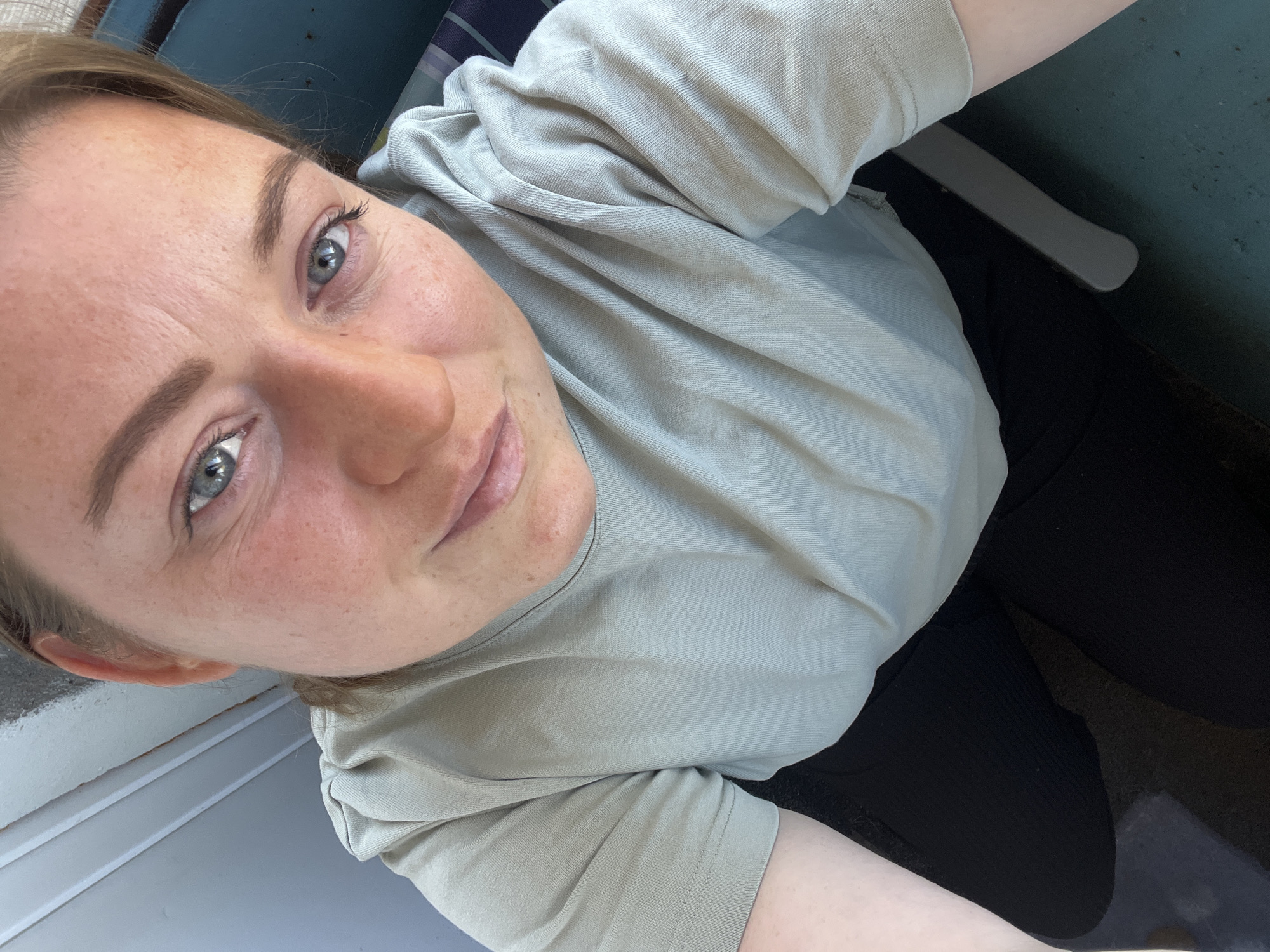 A selfie taken from a high angle shows a woman with fair skin, light brown hair, and blue eyes. She is wearing a light green T-shirt and black pants. The background includes a surface that appears to be painted blue and a white object, possibly part of a chair. The image does not provide specific information to confirm a location in the city of Раздолье, Russia.