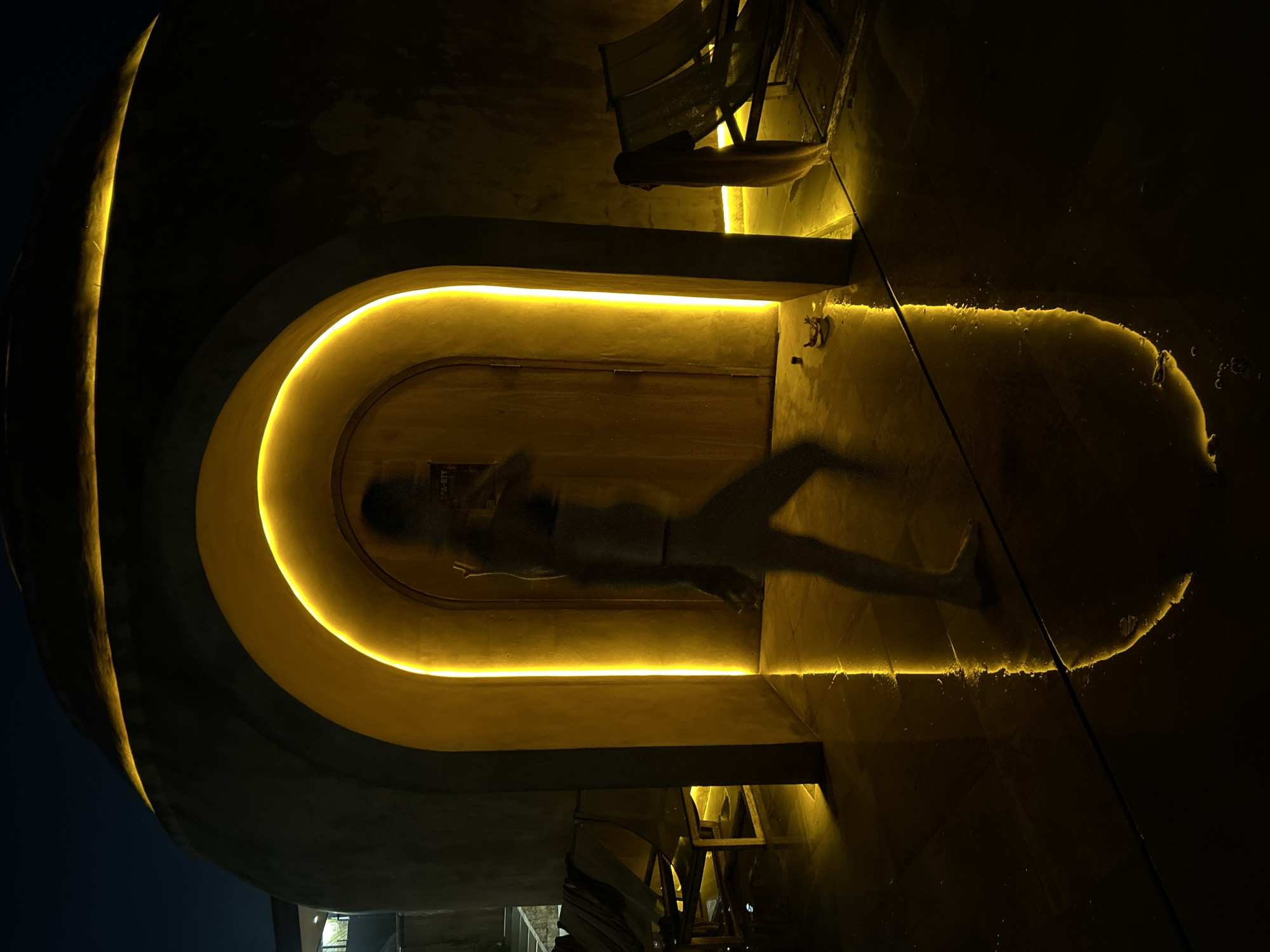 The image depicts a person, captured in motion blur, walking past an illuminated archway at night. The archway features a warm, yellow light that casts a strong reflection on the wet floor, creating a dramatic visual effect. The environment appears to be an outdoor space, possibly a resort or a spa, given the presence of lounge chairs and the overall ambiance. The dark sky and the artificial lighting suggest the photo was taken after sunset. The location is identified as Kediri, Indonesia, which implies a tropical or subtropical climate where such outdoor amenities might be common. The person in the image is silhouetted and indistinct due to the motion blur, but appears to be wearing swimwear. The scene is characterized by shadows, light reflections, and a sense of mystery.