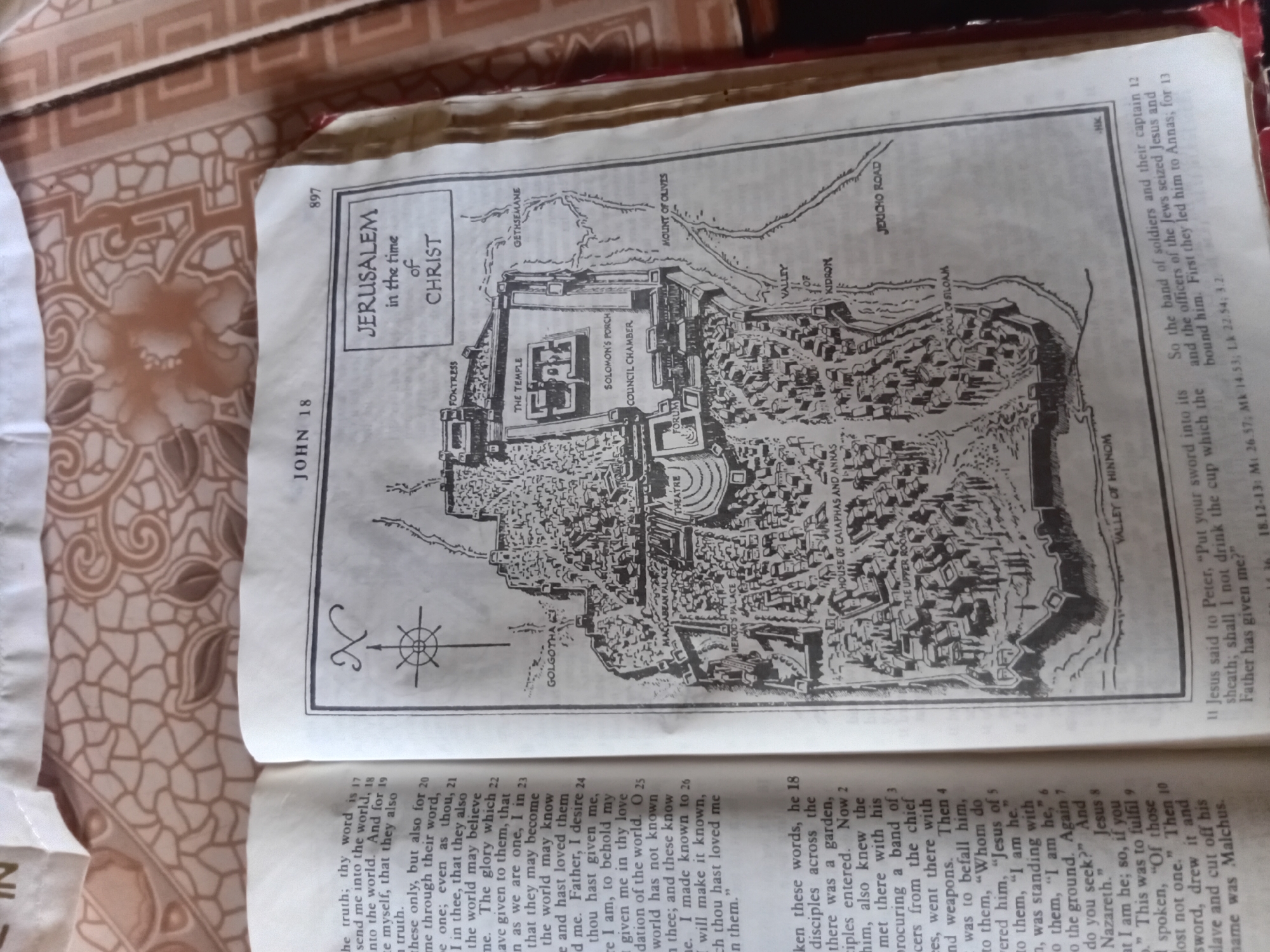 This media file is an image of an open book, lying on a brown and tan patterned fabric. The left page features a large, decorative letter "N" and a compass rose symbol, followed by text that appears to be scripture. The text is in English and includes verses from John 18.
The right page prominently displays a black and white illustration of "Jerusalem in the time of Christ." The map depicts the city with its walls, gates, and various significant locations labeled, such as "The Temple," "Solomon's Porch," "Council Chamber," "Mount of Olives," "Valley of Hinnom," and "Jericho Road." The map is detailed, showing buildings, streets, and geographical features. Above the map, there is a page number, "897," and the chapter heading, "JOHN 18." Below the map, more scripture text is visible, continuing from the left page and including references to Jesus' arrest and trial.
The overall impression is that of a page from a study Bible, providing a visual representation of a biblical setting to accompany the text. The lighting suggests an indoor environment.