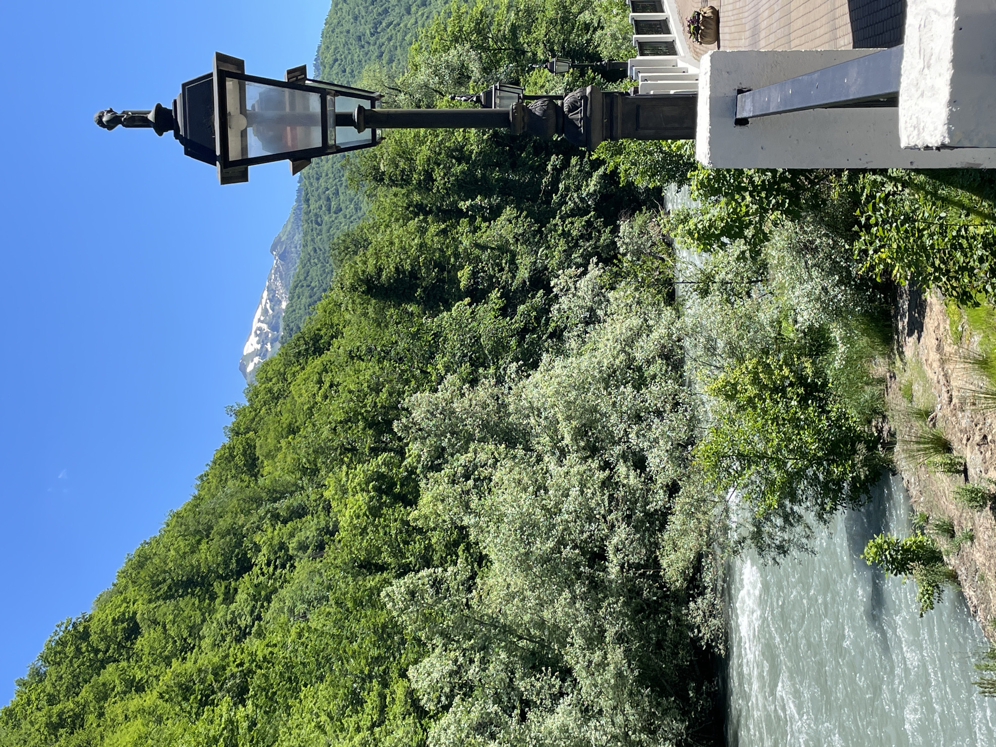 In Estosadok, Russia, a picturesque scene unfolds by a river under a clear blue sky. Lush green trees blanket the steep mountainside in the background, their vibrant foliage contrasting with the snowy peak visible in the distance. The river itself, a turbulent expanse of greyish-blue water, flows vigorously in the foreground. On the right side of the frame, a black ornate lamppost stands guard along a paved walkway bordered by a white railing. The walkway is constructed with light brown bricks and a small planter containing a flower adds a touch of color. The image captures the natural beauty of the landscape, with the man-made elements of the lamppost and walkway providing a sense of place within this serene environment.