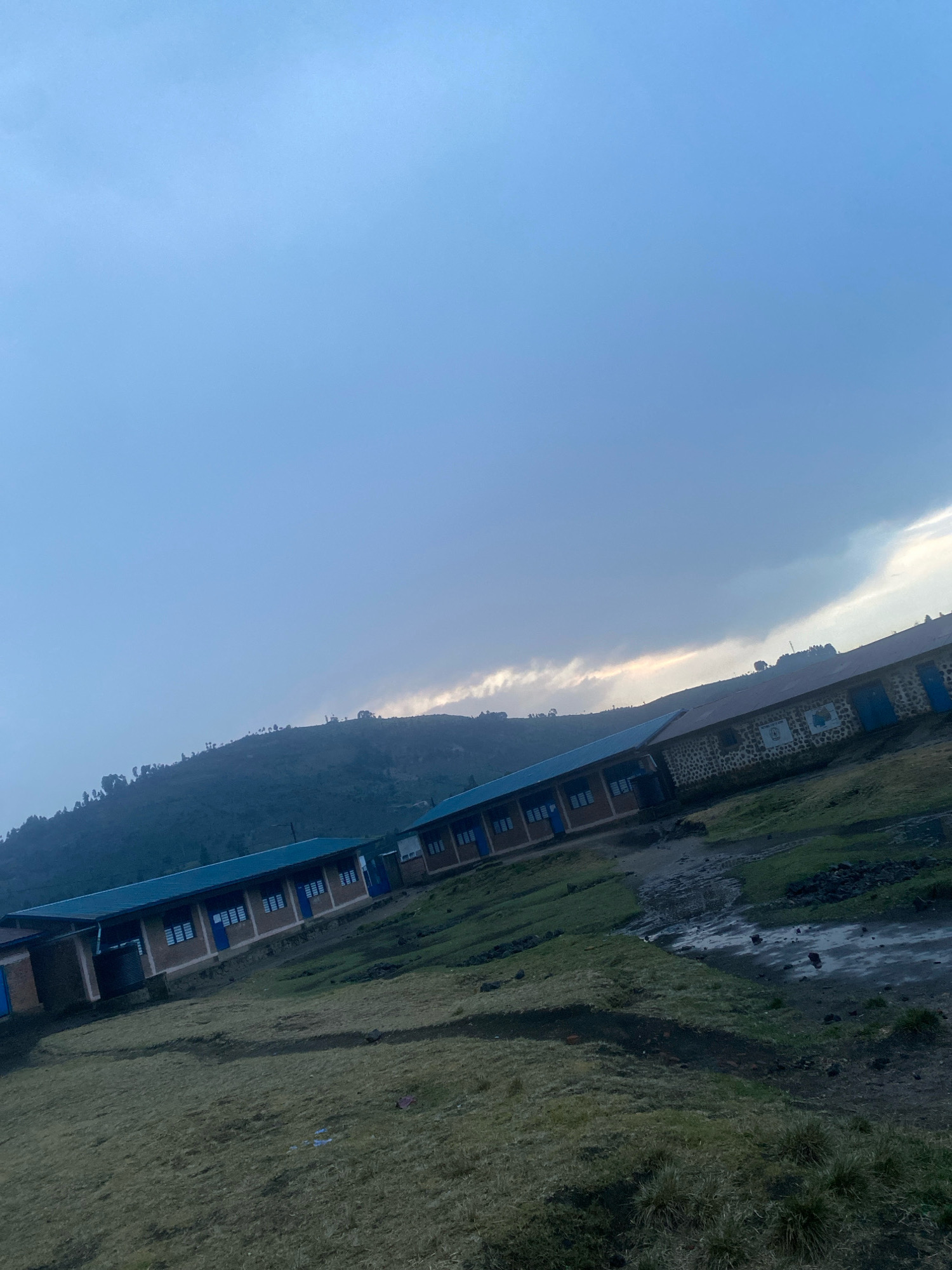 In Shaba, Rwanda, a photograph captures a rural school under a cloudy sky. The buildings, likely classrooms, are long and single-story with blue roofs and brick walls. Some walls are adorned with stone patterns and feature blue doors and windows. The foreground shows a grassy, uneven terrain with patches of mud and scattered rocks. In the background, a lush, green hillside slopes upwards, dotted with trees and the silhouette of a distant structure. The overall scene suggests a serene, remote educational setting.