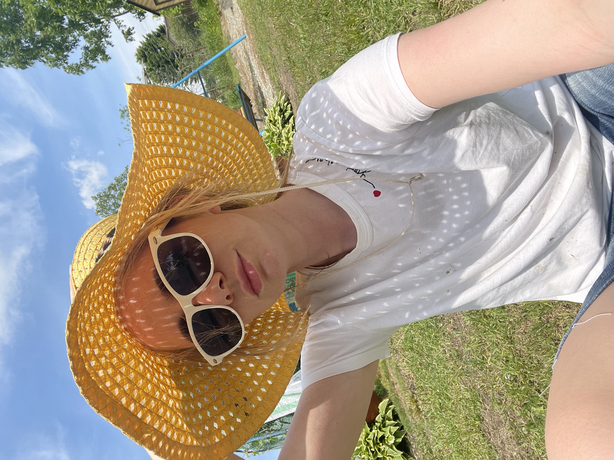 A person wearing a wide-brimmed yellow straw hat and white-framed sunglasses poses in a grassy outdoor setting. They are also wearing a white t-shirt with a small graphic on the chest, and blue denim bottoms are partially visible. The sky is blue with some clouds in the background, and there is greenery including grass and plants in the frame. The location is potentially in Yasnoe, Russia, though there are no specific elements to definitively identify it as such.