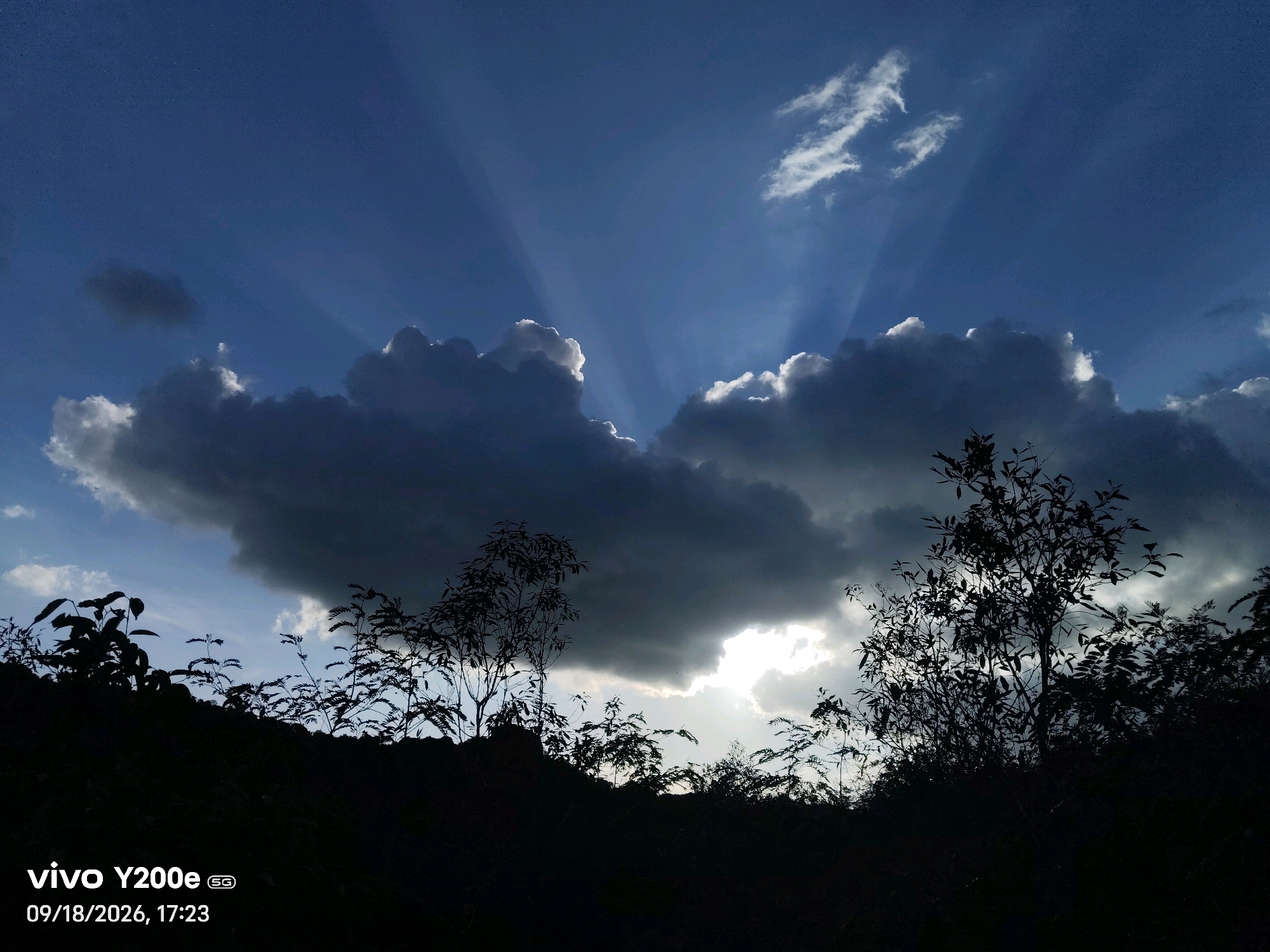 The image captures a dramatic sky over the Tippagondanahalli reservoir in India, on September 18, 2026, at 5:23 PM. The sun is setting, and its rays are piercing through dark, heavy clouds, creating a crepuscular effect. These beams of light fan out across the deep blue sky, illuminating the edges of the clouds with a bright white glow. In the foreground, the silhouette of the landscape is visible, with dark trees and vegetation forming a stark contrast against the bright sky. The terrain appears to be a gentle slope leading towards the reservoir, which is not directly visible in this frame. The overall atmosphere is one of impending twilight, with a sense of natural beauty and a touch of drama due to the weather conditions. The timestamp and device information "vivo Y200e 5G" are present in the lower-left corner of the image.