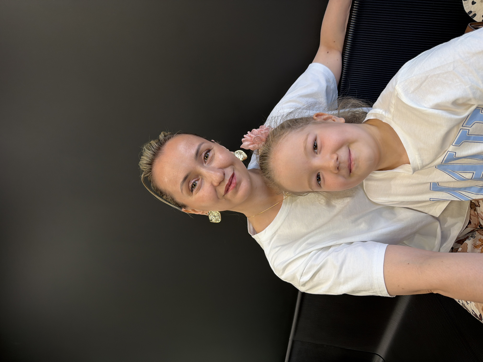 A woman and a young girl are smiling and posing closely together for a photo. The woman has blonde hair pulled back with a headband, a gold necklace, and gold earrings. She is wearing a white t-shirt. The girl has blonde hair and a pink flower clip in her hair. She is also wearing a white t-shirt with light blue writing. They are both facing the camera. The background is primarily a dark gray color, with a black textured surface visible on the right side. The location is unknown and cannot be confirmed to be Izhevsk, Russia based on the image.