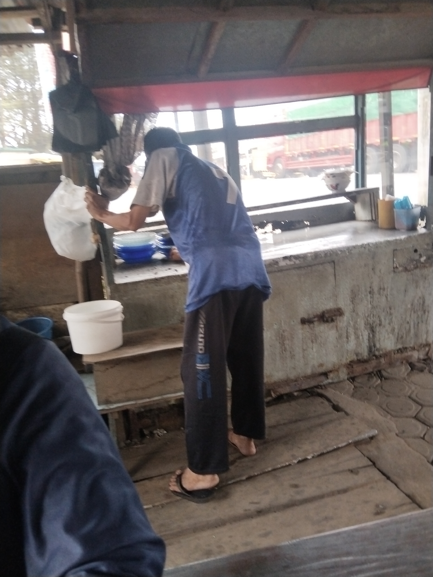 In the city of Tanjung Marwo, Indonesia, a person stands on a wooden platform, facing away from the camera. They are wearing a blue and grey shirt, black pants with a white stripe, and black flip-flops. The person is holding onto a pole with a plastic bag hanging from it. To the left of the person is a white bucket, and to the right are stacked blue and white plates. Behind the person is a counter with various items, including a bowl with a pattern, and yellow and blue containers. Through a window behind the counter, a red truck with a green load is visible. The environment appears to be an outdoor market or food stall, with a rustic wooden structure overhead and a tiled floor beyond the platform. The overall scene suggests a commercial area, possibly a food vendor's stall.