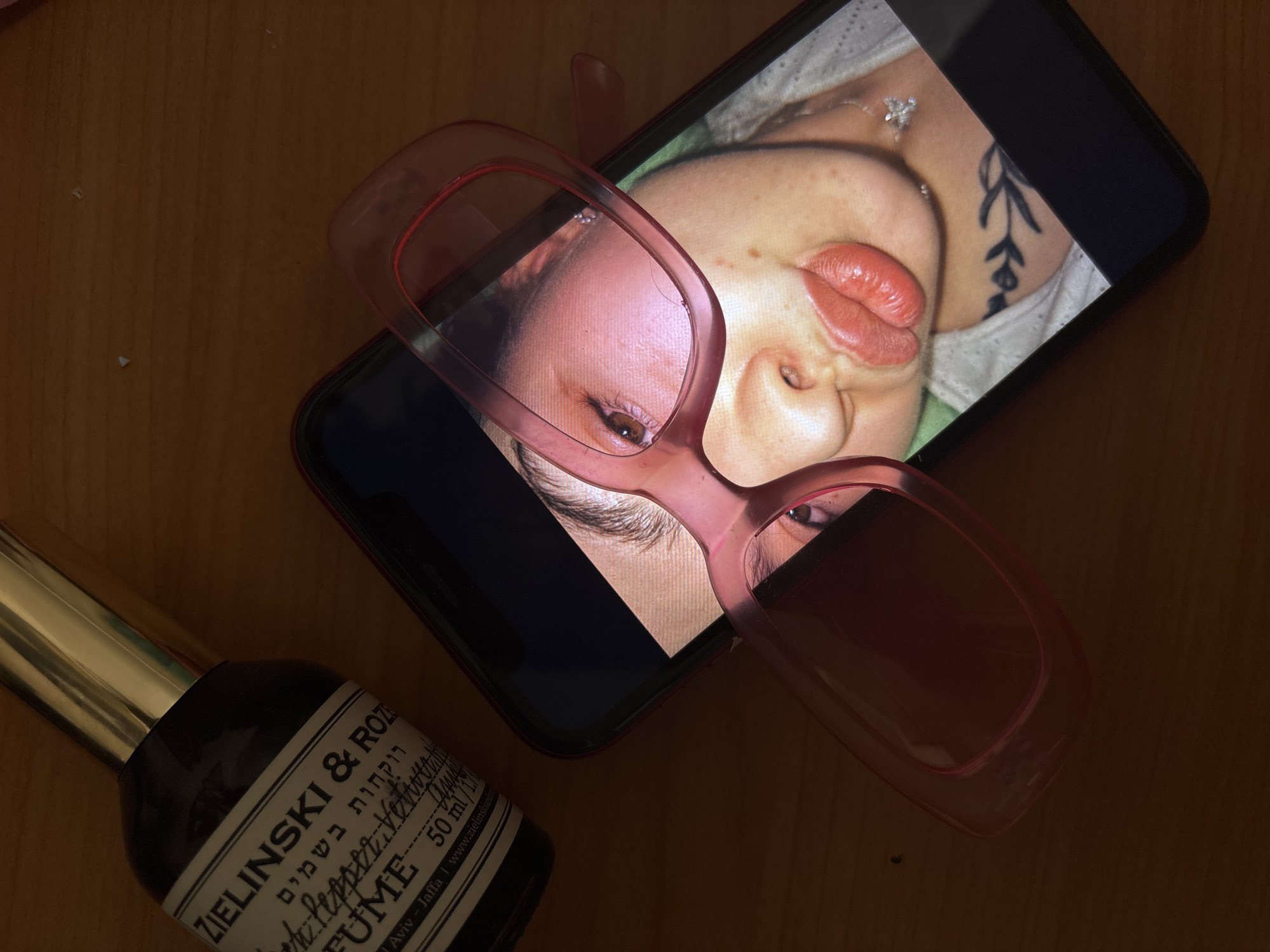 The image displays a flat lay composition of personal items on a dark, wooden surface. A smartphone, oriented lengthwise, presents a close-up image of a woman's face, visible sideways and inverted on the screen. Her features include full, red lips, a small nose piercing, a portion of her brown eye, and a tattoo of a leafy branch on her upper left chest or shoulder. She wears a delicate necklace with a small pendant and a light-colored, possibly lace-trimmed garment.
Resting partially on the smartphone screen are pink-tinted, clear-framed glasses. One lens of the glasses refracts and magnifies a portion of the woman's eye from the screen. To the lower left, a dark glass perfume bottle with a gleaming gold-colored cap is positioned. The bottle's label, featuring both English and Hebrew text, is partially visible. Identifiable English text includes "ZIELINSKI & ROZEN", "PERFUME", "Black Pepper, Vetiver", "50 ml", and "Tel Aviv - Jaffa | www.zielinskiande". The Hebrew text "רוקחות בשמים" (Perfumery) is also visible. The overall lighting is dim, indicating an indoor setting, likely in the evening or a dimly lit room. The scene is situated in Perm, Russia.