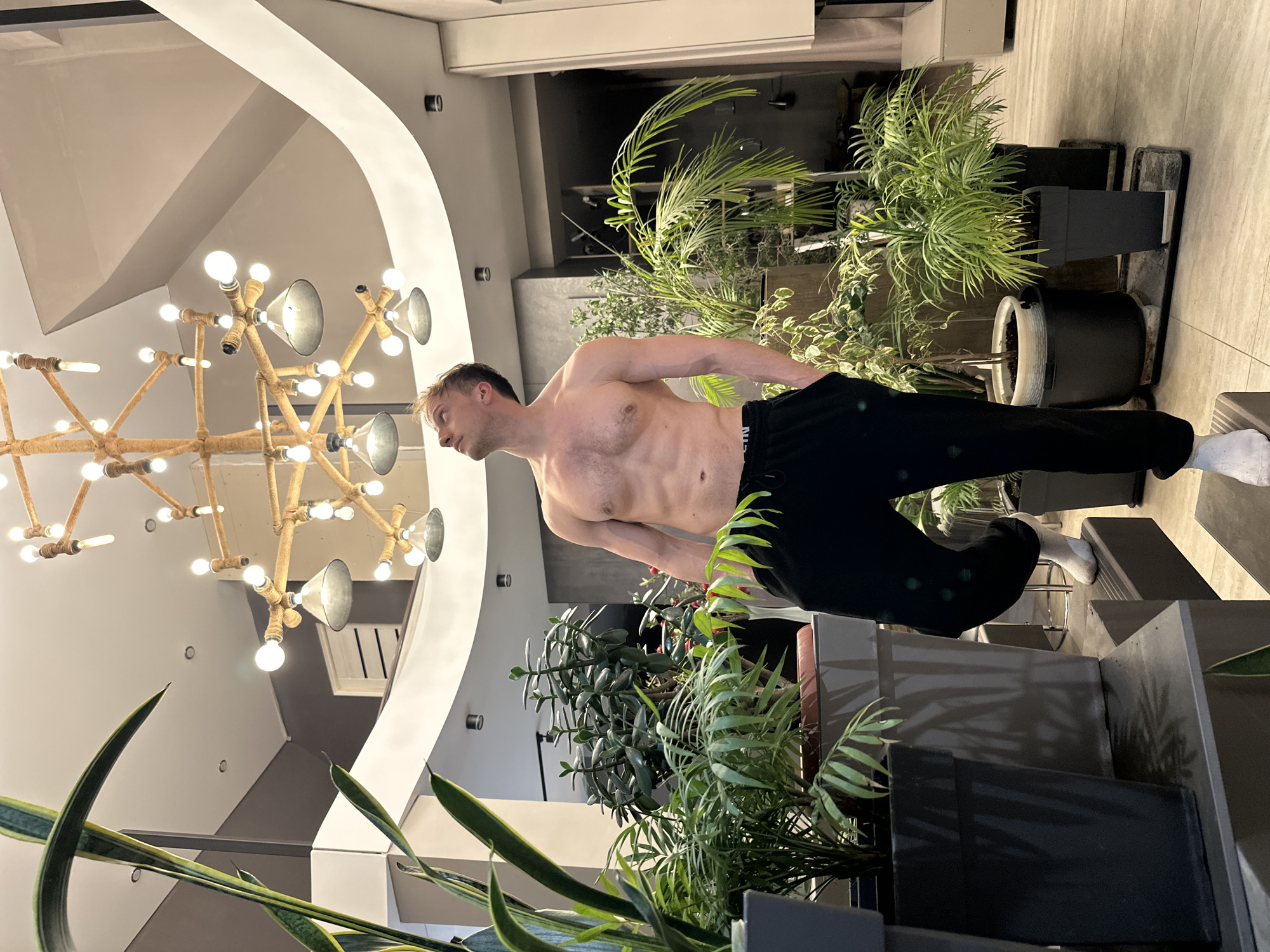 A shirtless man wearing black pants and white socks stands in a room with several potted plants. A large chandelier hangs from the ceiling. The location is Kursk, Russia.