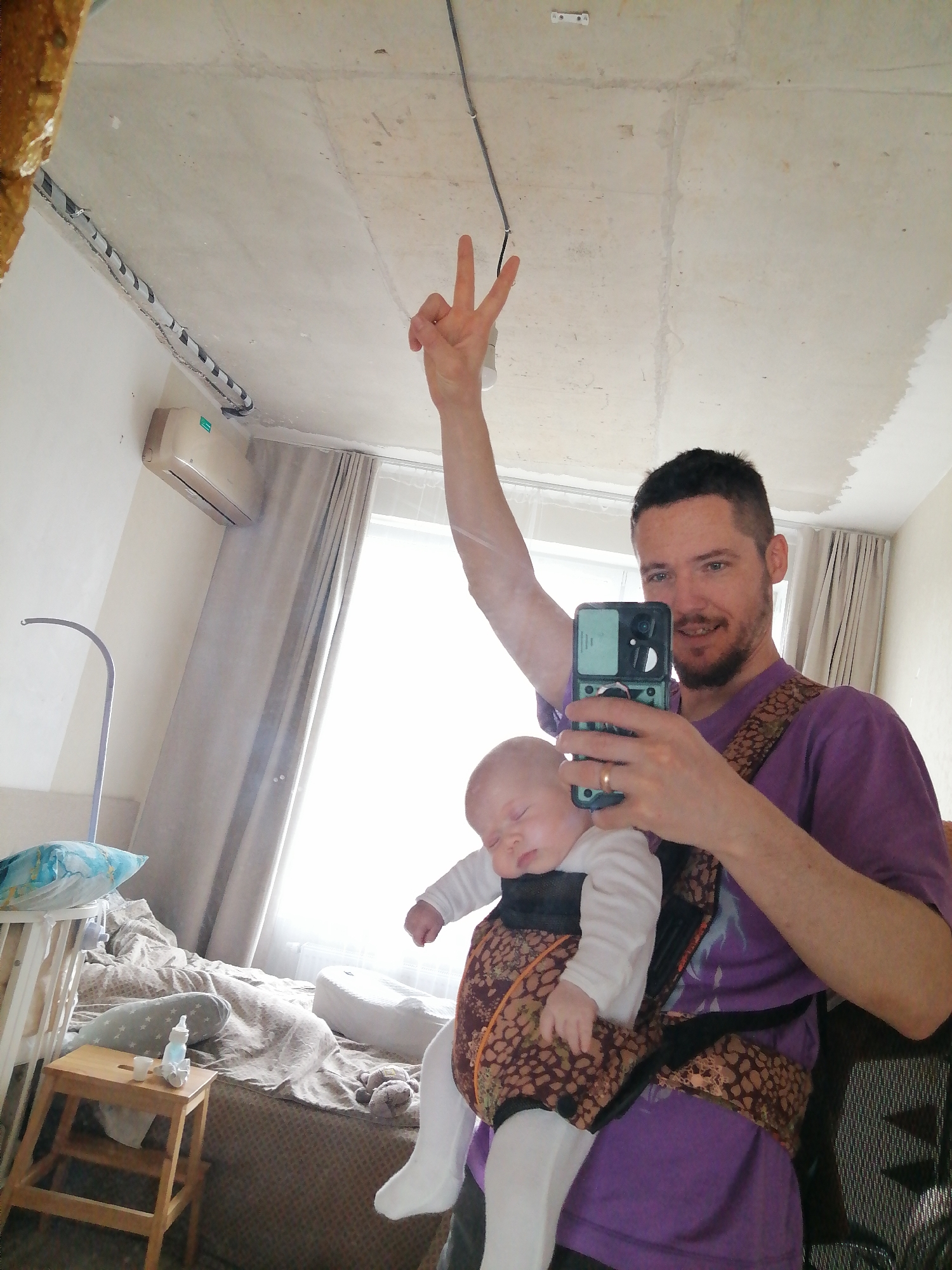 A man is taking a selfie with a baby in a carrier. The man is wearing a purple t-shirt and holding a phone. He is making a peace sign with his right hand. The baby is wearing a white shirt and white tights. The room appears to be a bedroom, with a crib, a bedside table, and a bed visible in the background. An air conditioning unit is mounted on the wall. The location appears to be a residential setting of Unknown, Unknown.