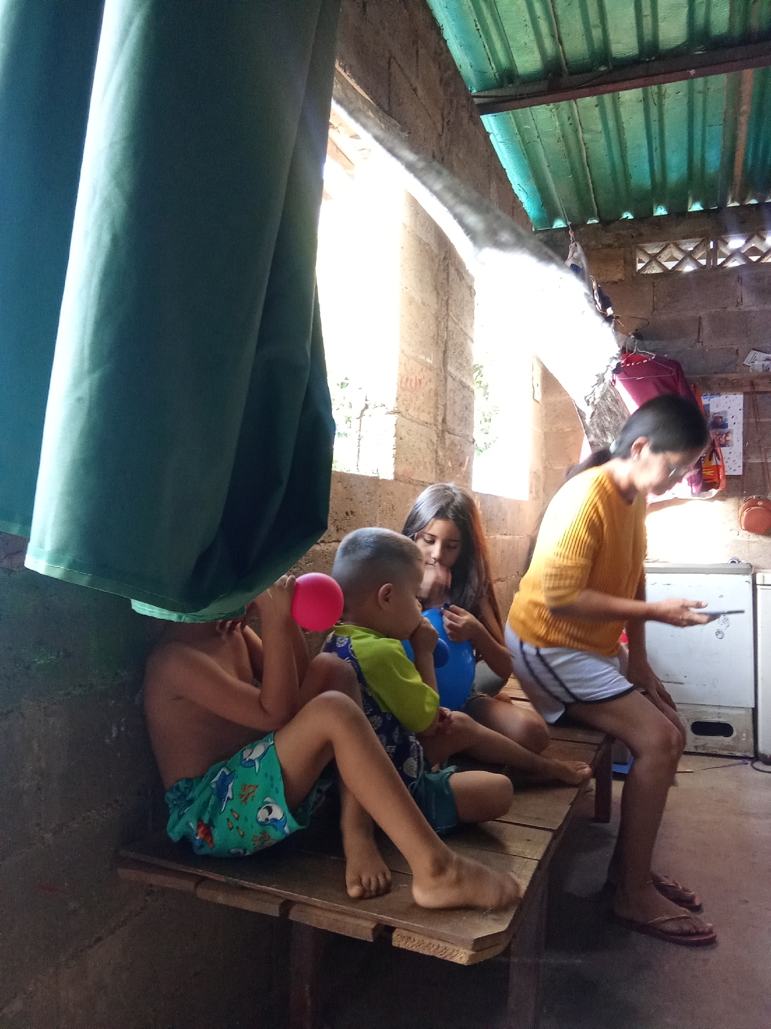 Four children are sitting on a wooden bench inside a dimly lit, sparsely furnished room. A large green curtain hangs to the left. A child on the left is blowing up a pink balloon. A child next to them is blowing up a blue balloon. A girl sits next to them. A woman wearing an orange shirt and white shorts sits on the edge of the bench looking at a mobile phone. Behind her are brick walls and a corrugated metal roof. A small refrigerator and a calendar are visible. The scene appears to be a house in Juan Griego, Venezuela.